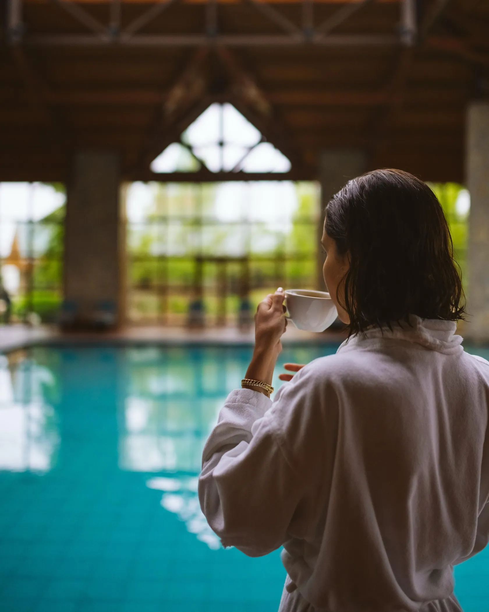A person in a robe holds a cup near an indoor pool under a wooden structure.