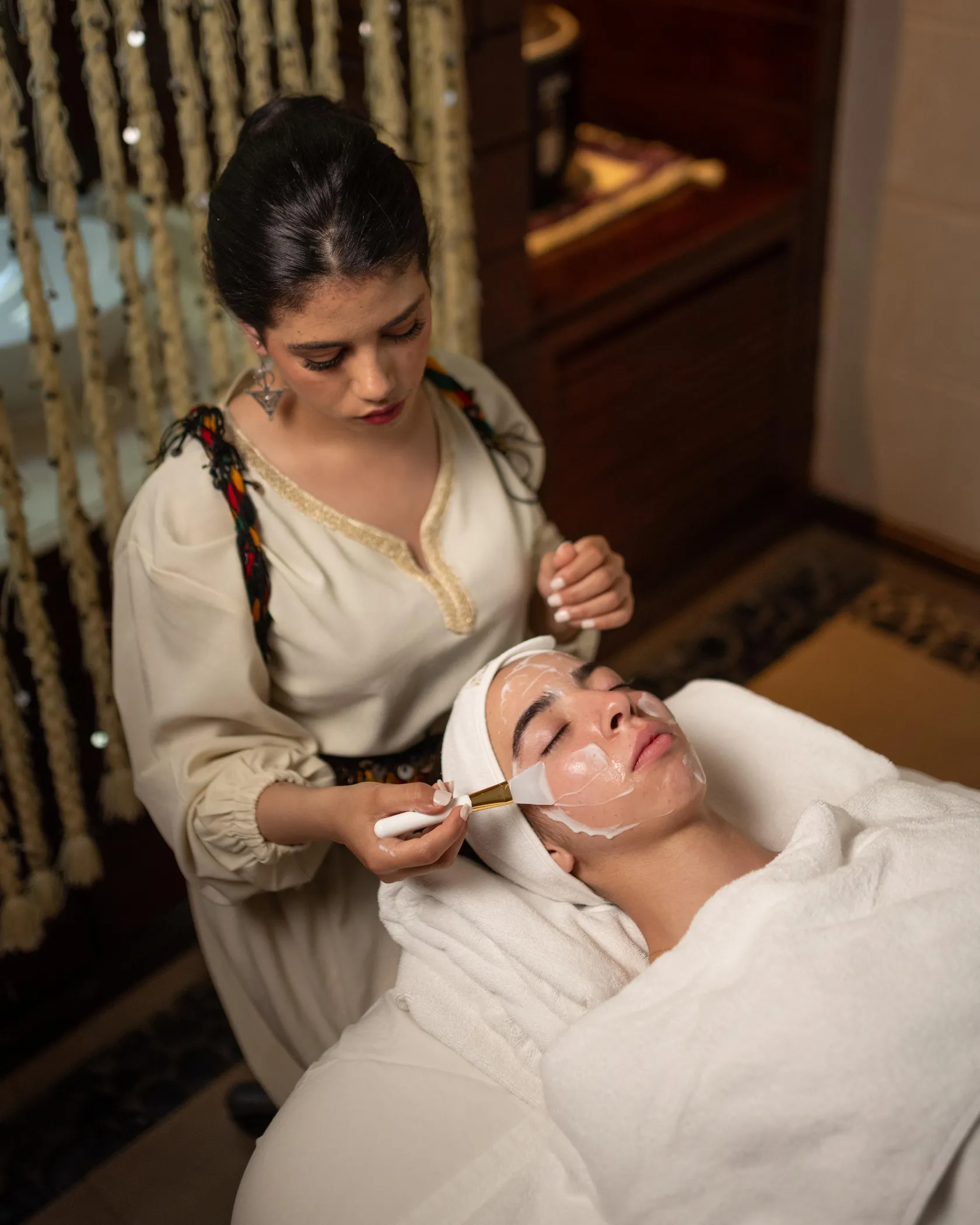 A person applies a facial mask to another person lying on a white towel.