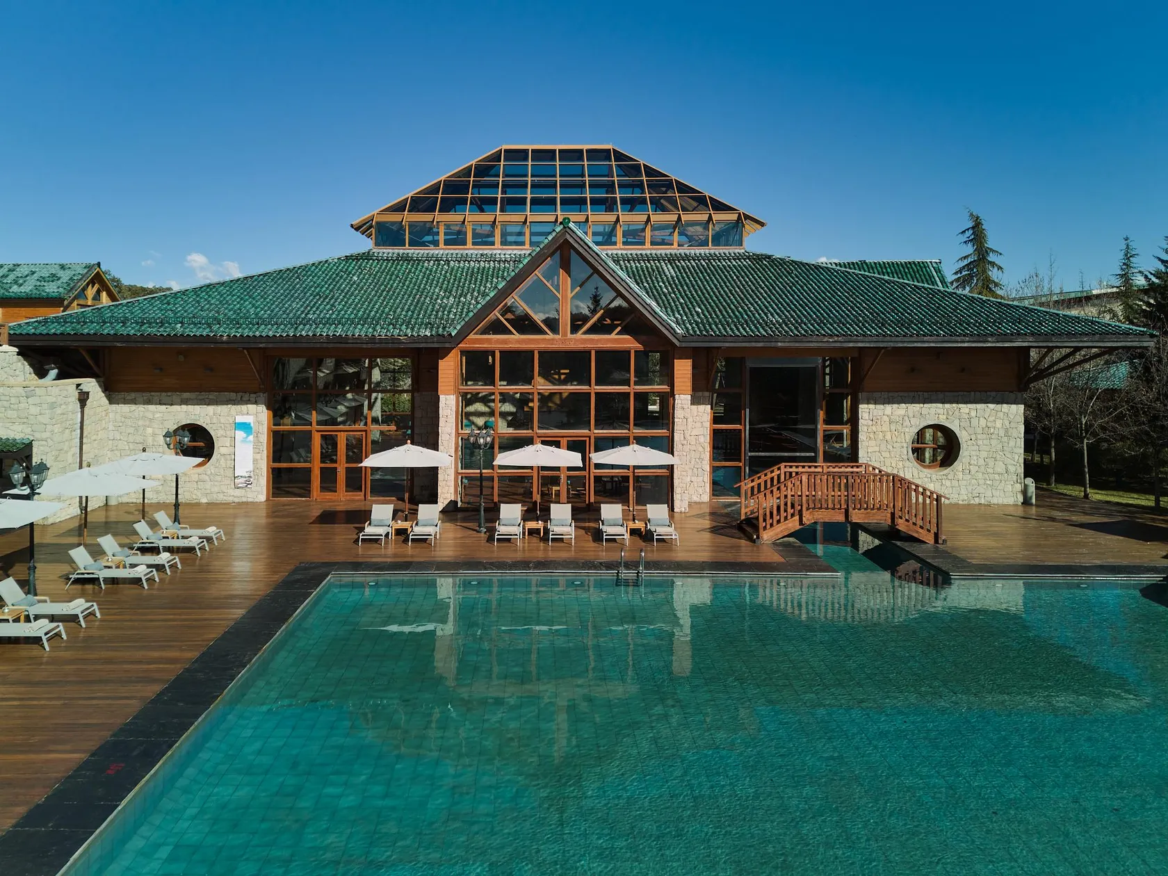 A stone and wood building with a green roof and a glass extension, in front of an outdoor pool surrounded by lounge chairs and umbrellas.