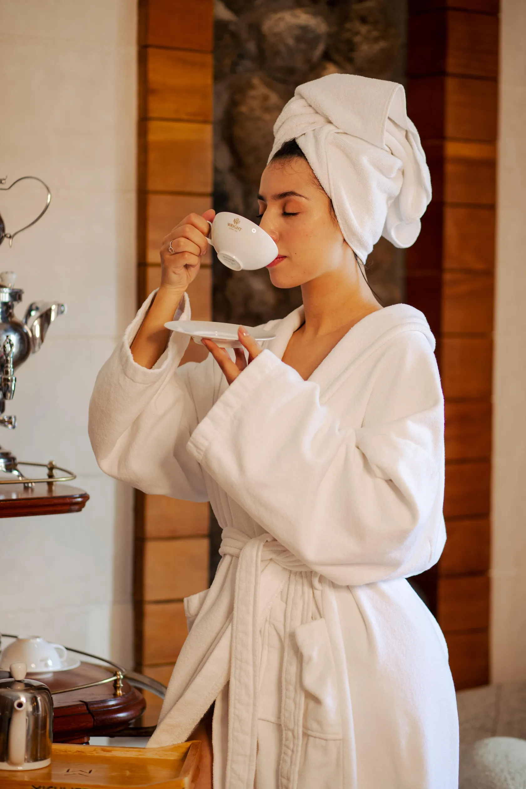 A person in a white bathing robe drinks a hot beverage from a white cup, with a towel wrapped around their head, in front of a mirror and bathroom accessories.