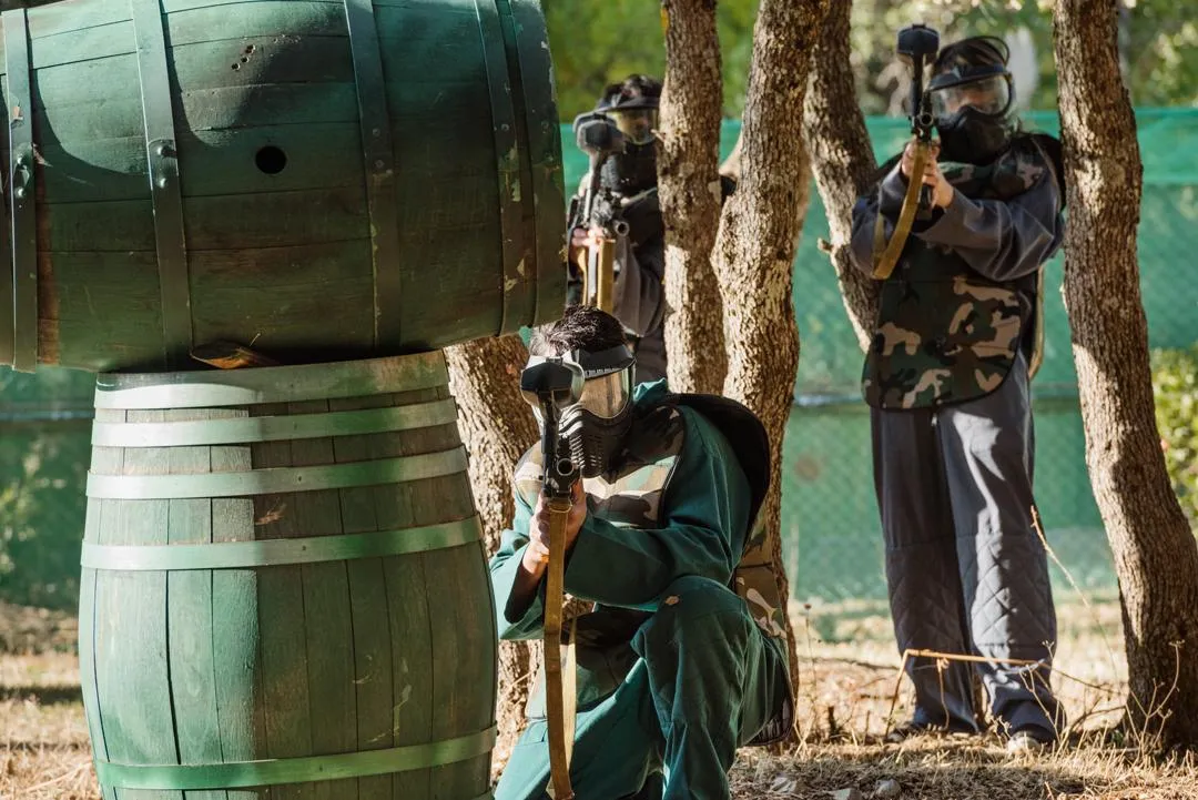 Three people in camouflage clothing and protective masks are hiding behind wooden barrels and trees, holding fake weapons.