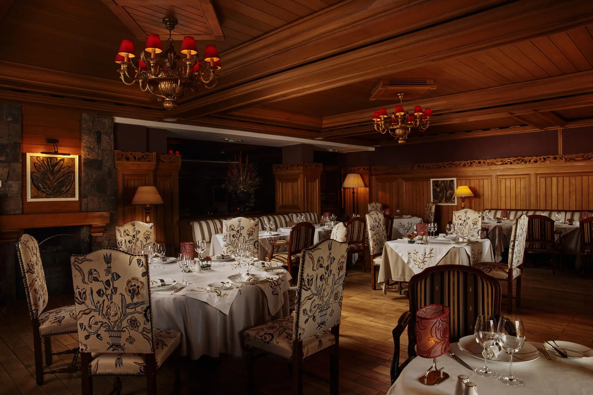 Elegant restaurant with set tables, chandeliers, and floral-patterned chairs, lit by lamps and chandeliers