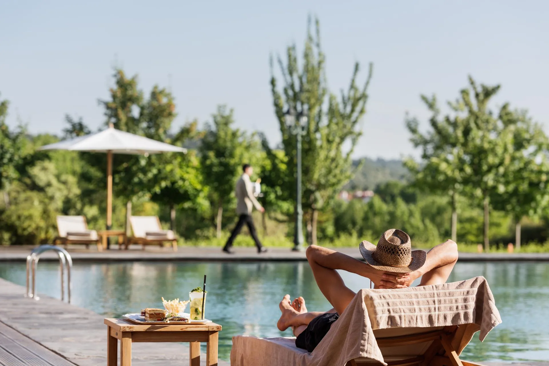 Une personne en maillot de bain et chapeau repose sur une chaise longue au bord d'une piscine, avec une table basse contenant des boissons et de la nourriture à proximité
