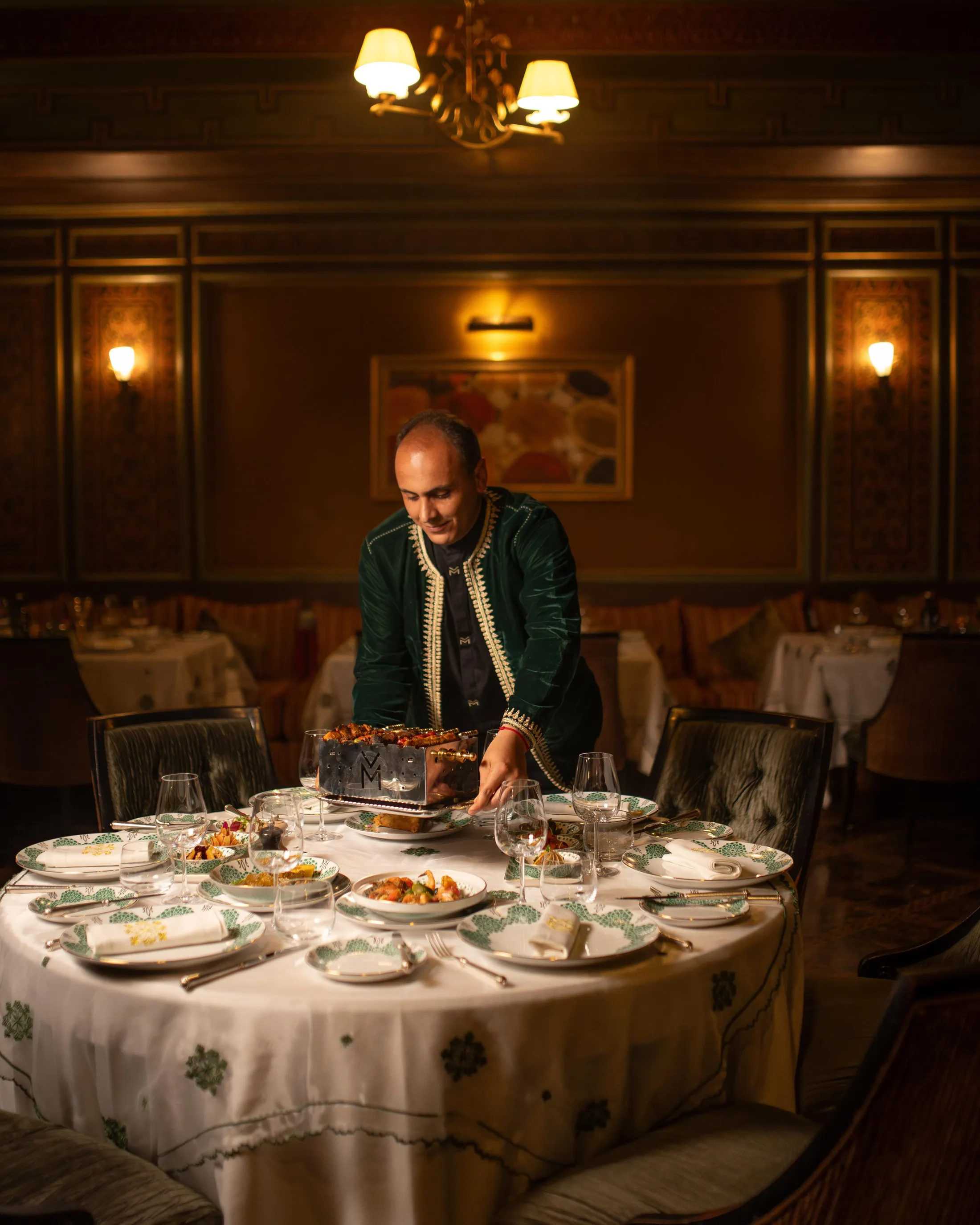 A man in a green jacket serves a dish at a formal dinner in an elegant dining room with chandeliers and decorated walls.