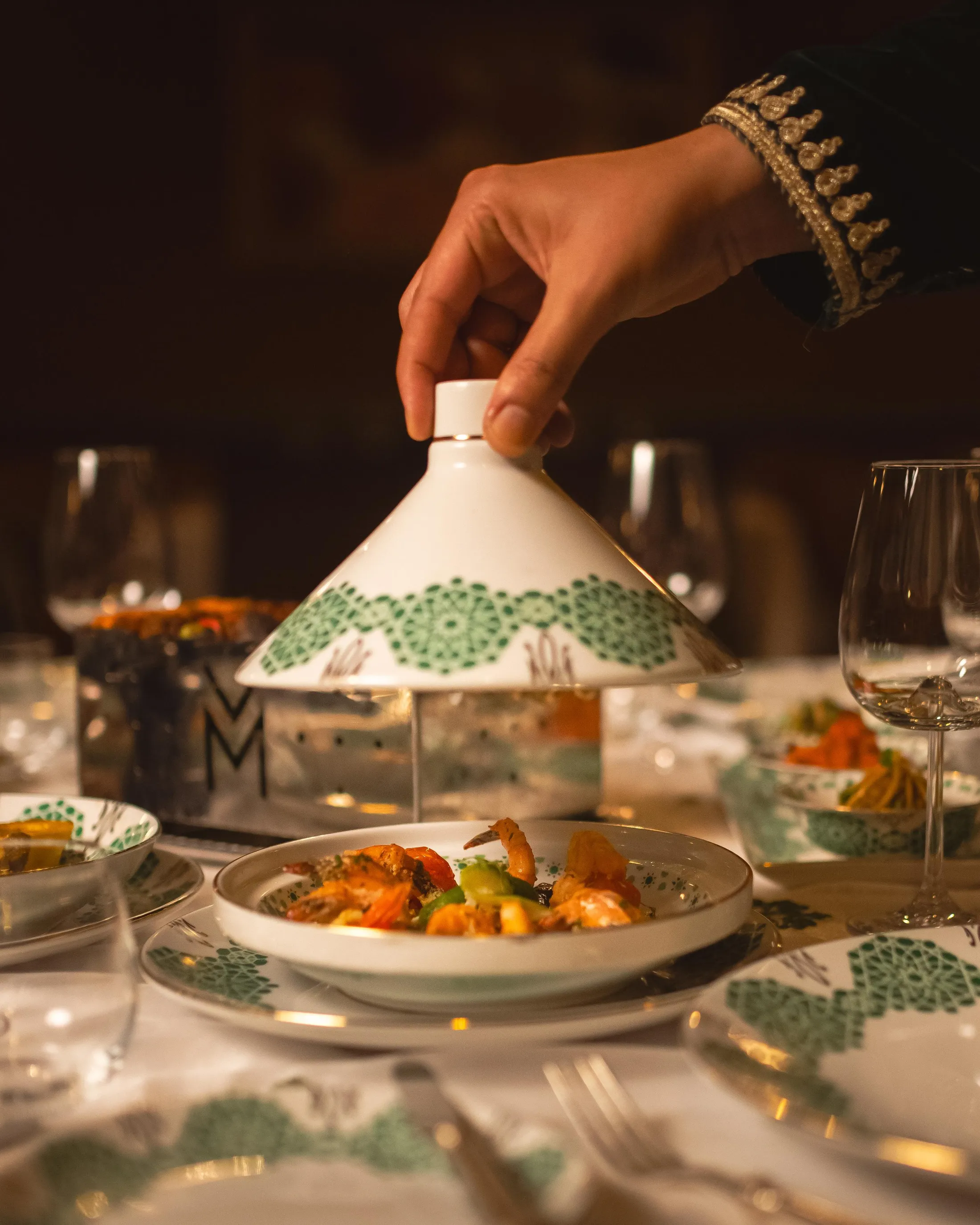 Une main tenant un couvercle en céramique au-dessus d'un plat de nourriture sur une table dressée
