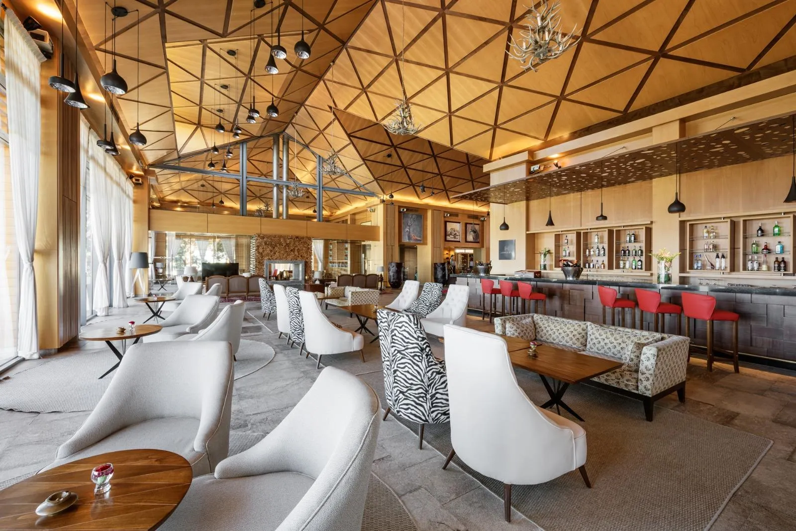 Un salon élégant avec des canapés, des chaises et des tables basses, éclairé par des suspensions et des lampes murales, avec un bar et des étagères à bouteilles en arrière-plan