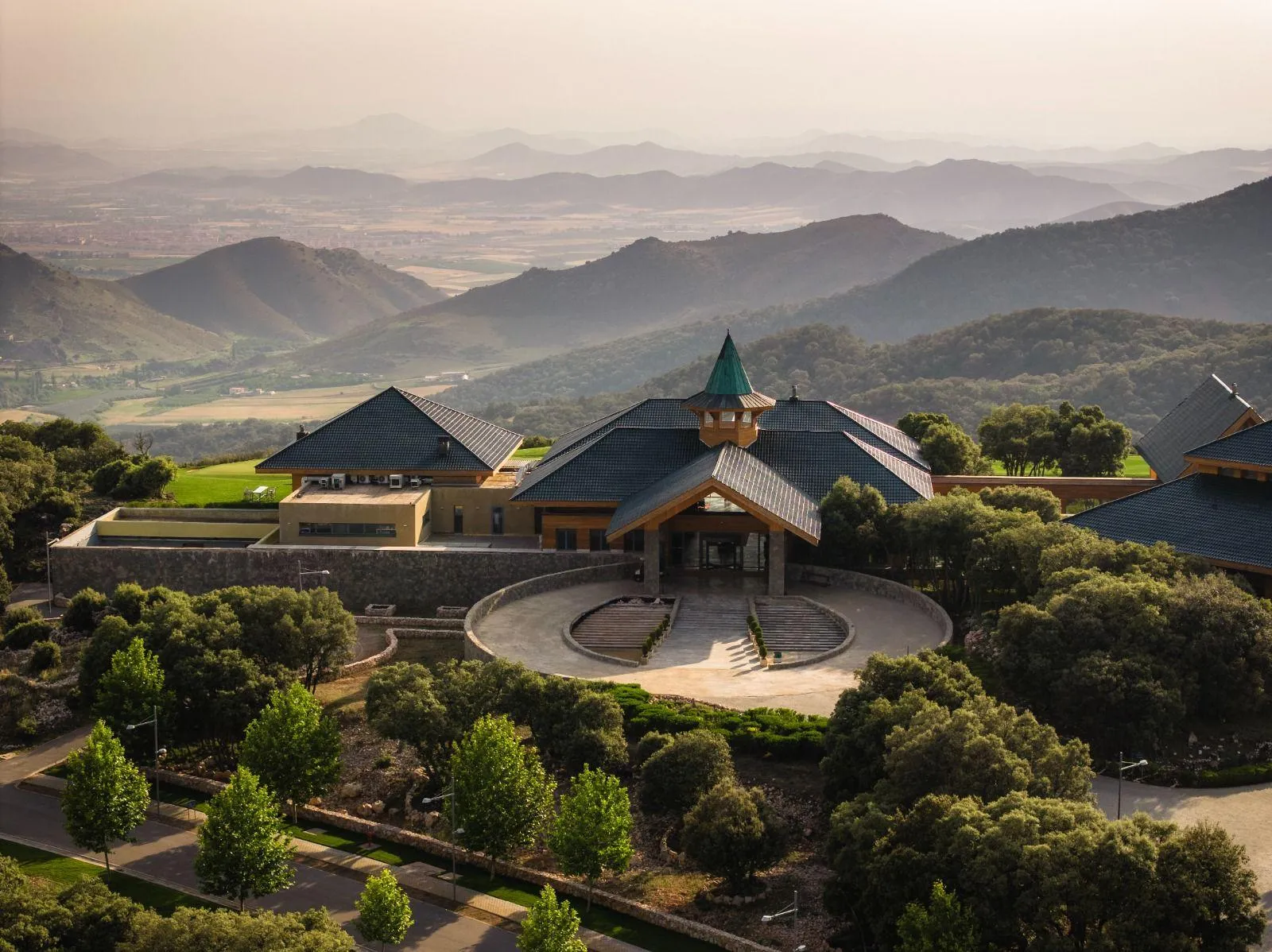 A building with steep roofs and a central tower stands in a rolling, green landscape, surrounded by trees and gardens.