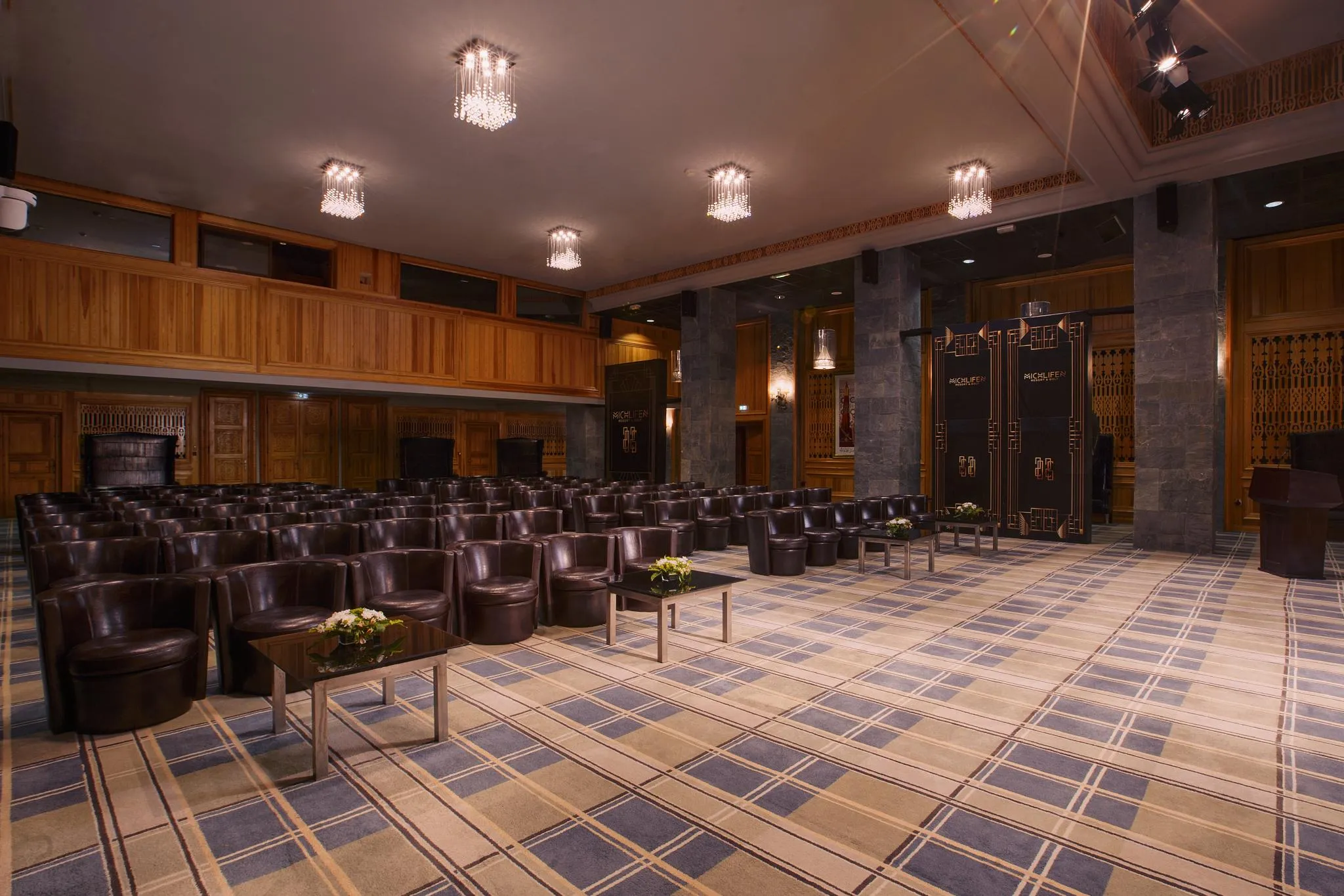 Conference room with rows of brown leather armchairs, low tables with flowers, wooden walls, and chandeliers on the ceiling.
