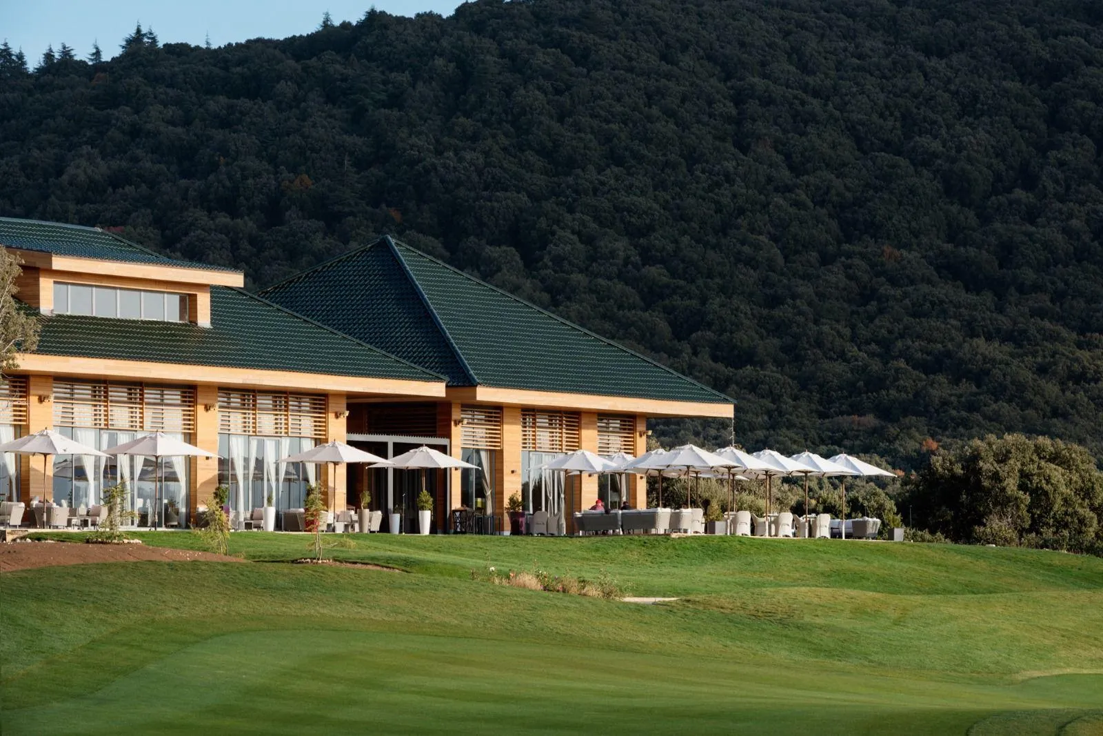 A building with a green roof and outdoor terraces equipped with tables and chairs under umbrellas, located near a golf course with a wooded hill in the background.
