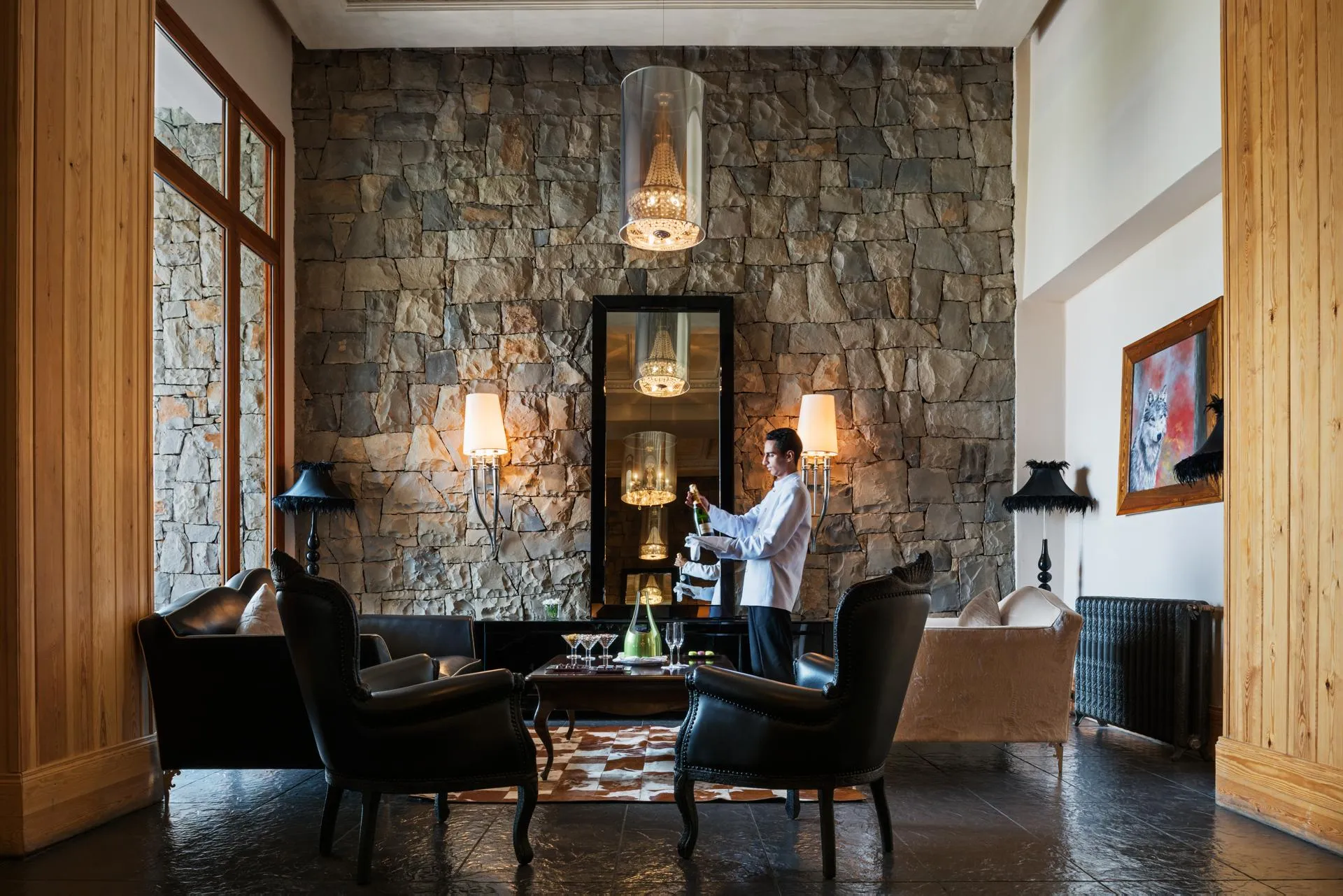 A waiter in a white jacket prepares drinks behind a stone bar with candelabras, surrounded by armchairs and lamps in an elegant lounge.