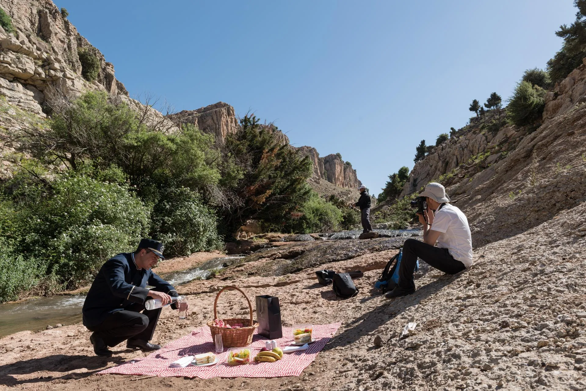Two people are having a picnic near a stream in a rocky canyon, with fruit, a bottle, and a basket on a blanket.