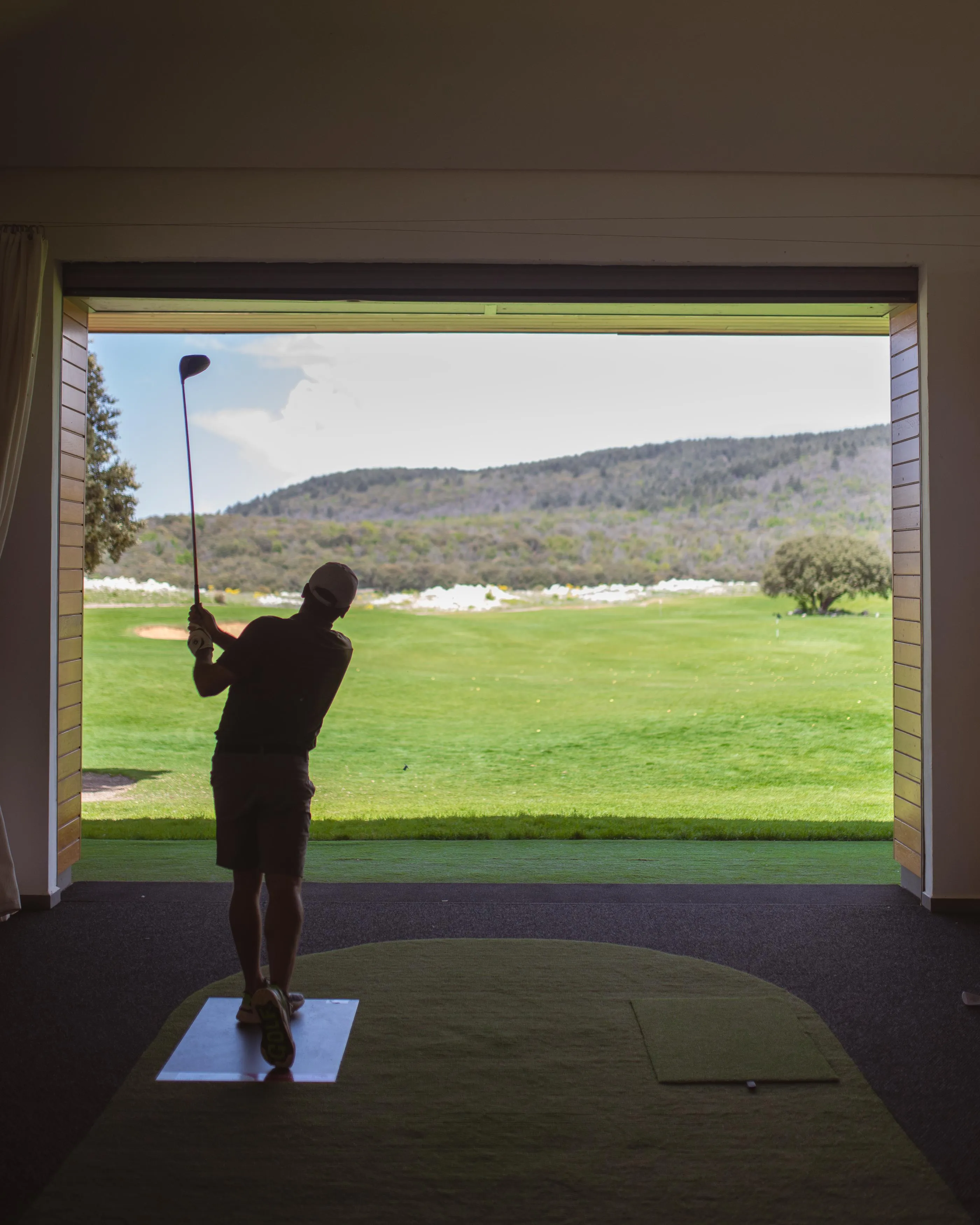 Silhouette of a person playing golf inside a building with an opening overlooking an outdoor golf course
