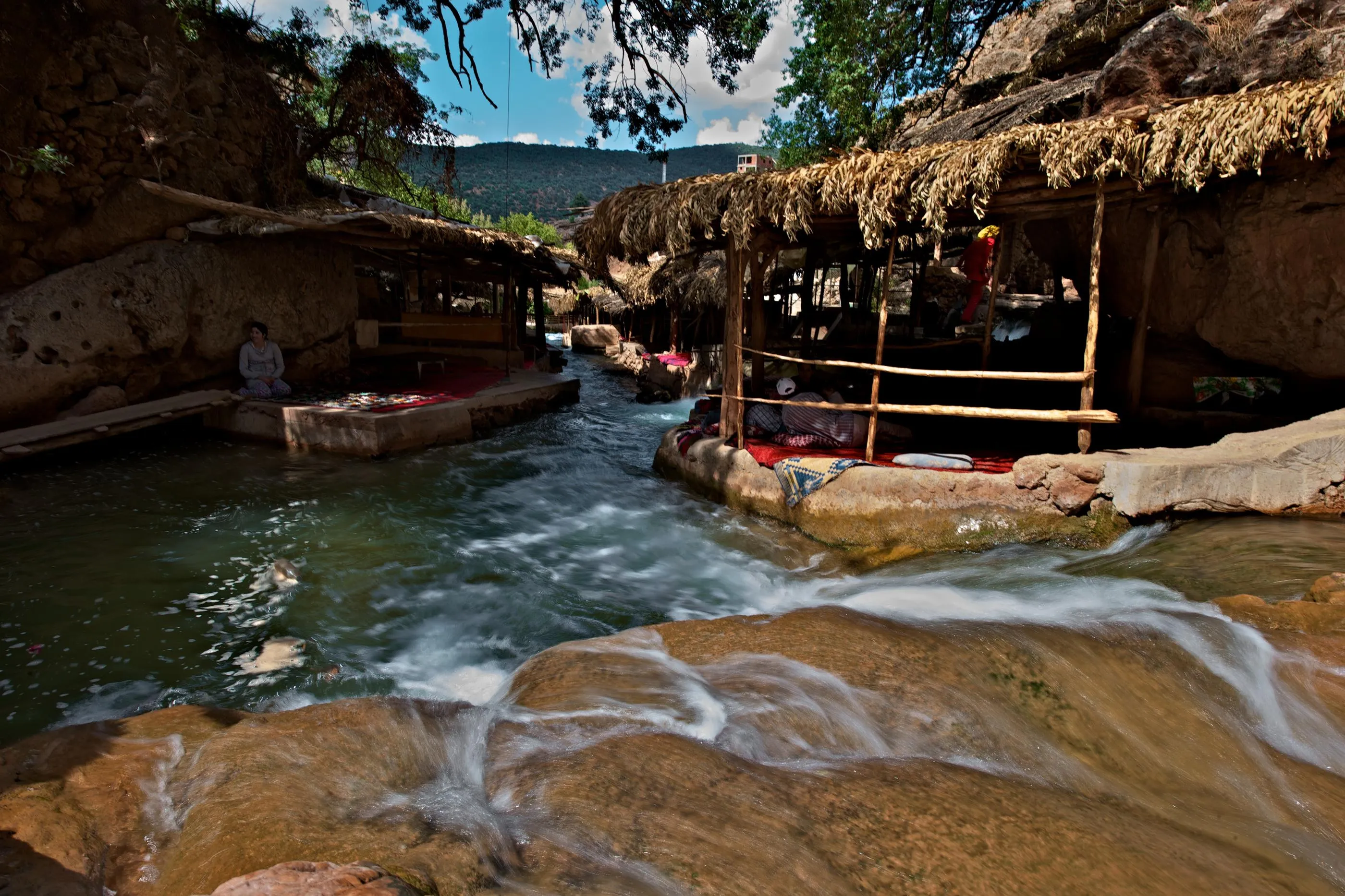 A watercourse with people sitting on stone platforms under thatched shelters