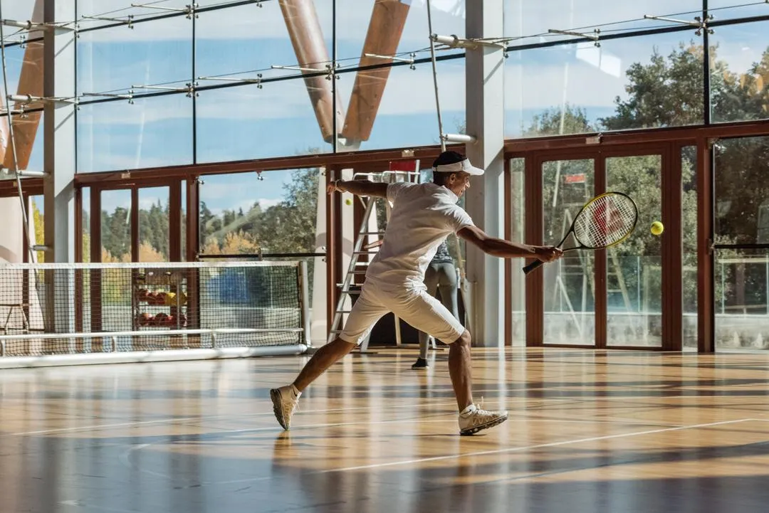 A man is playing tennis in an indoor hall with large glass windows, hitting a ball with his racket.