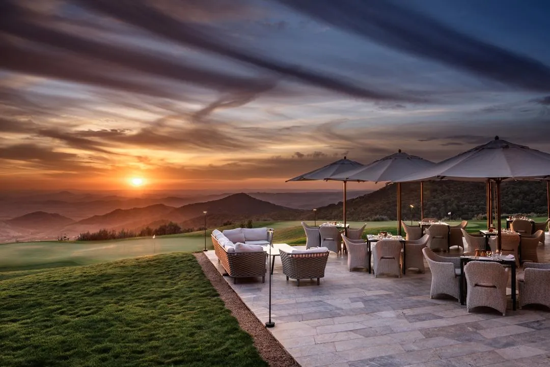 Terrace with chairs and tables under umbrellas, facing a sunset over mountains and a golf course.