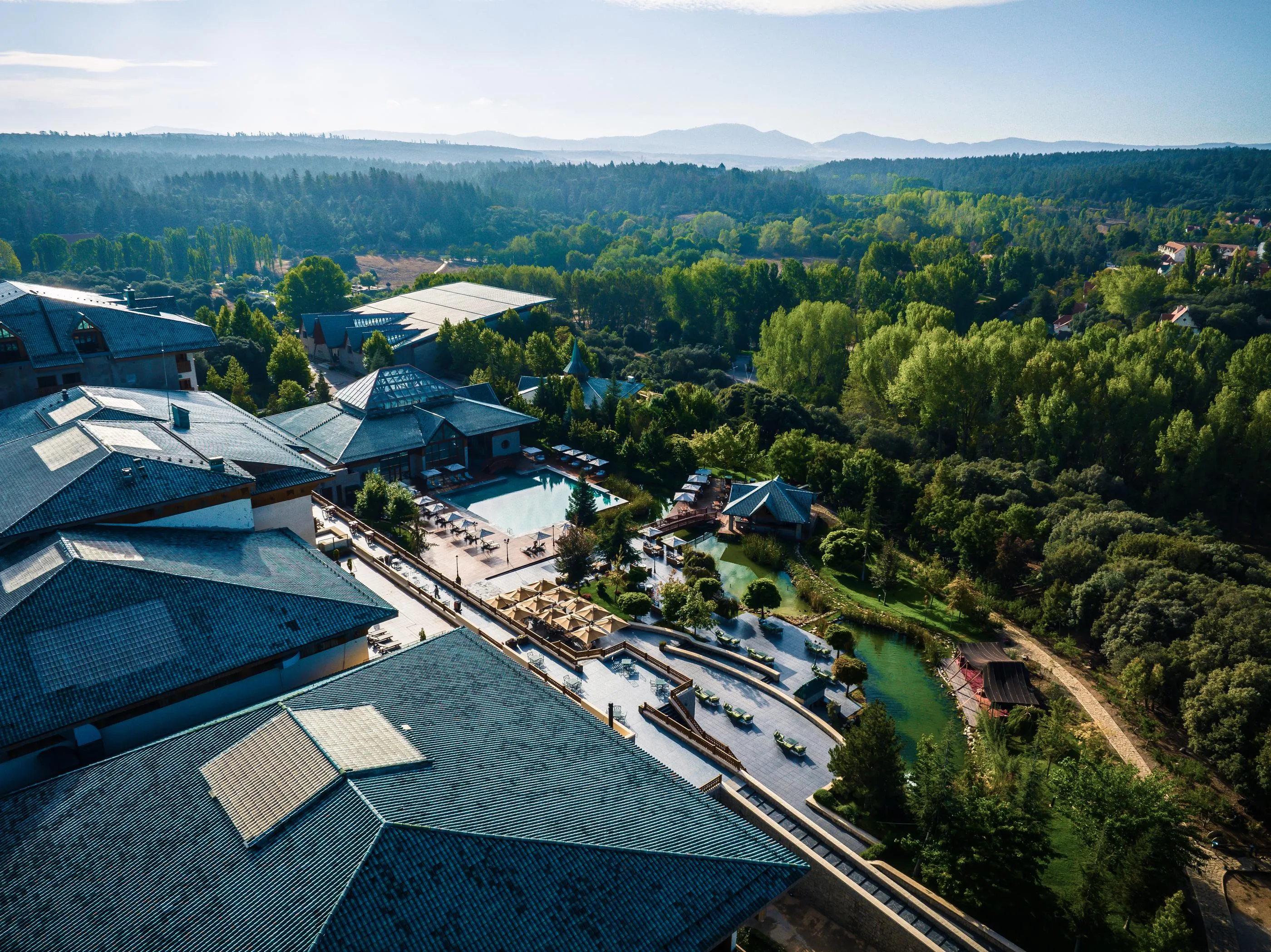 Aerial view of a complex of traditional buildings with tiled roofs, surrounded by trees and water bodies, in a mountainous landscape.