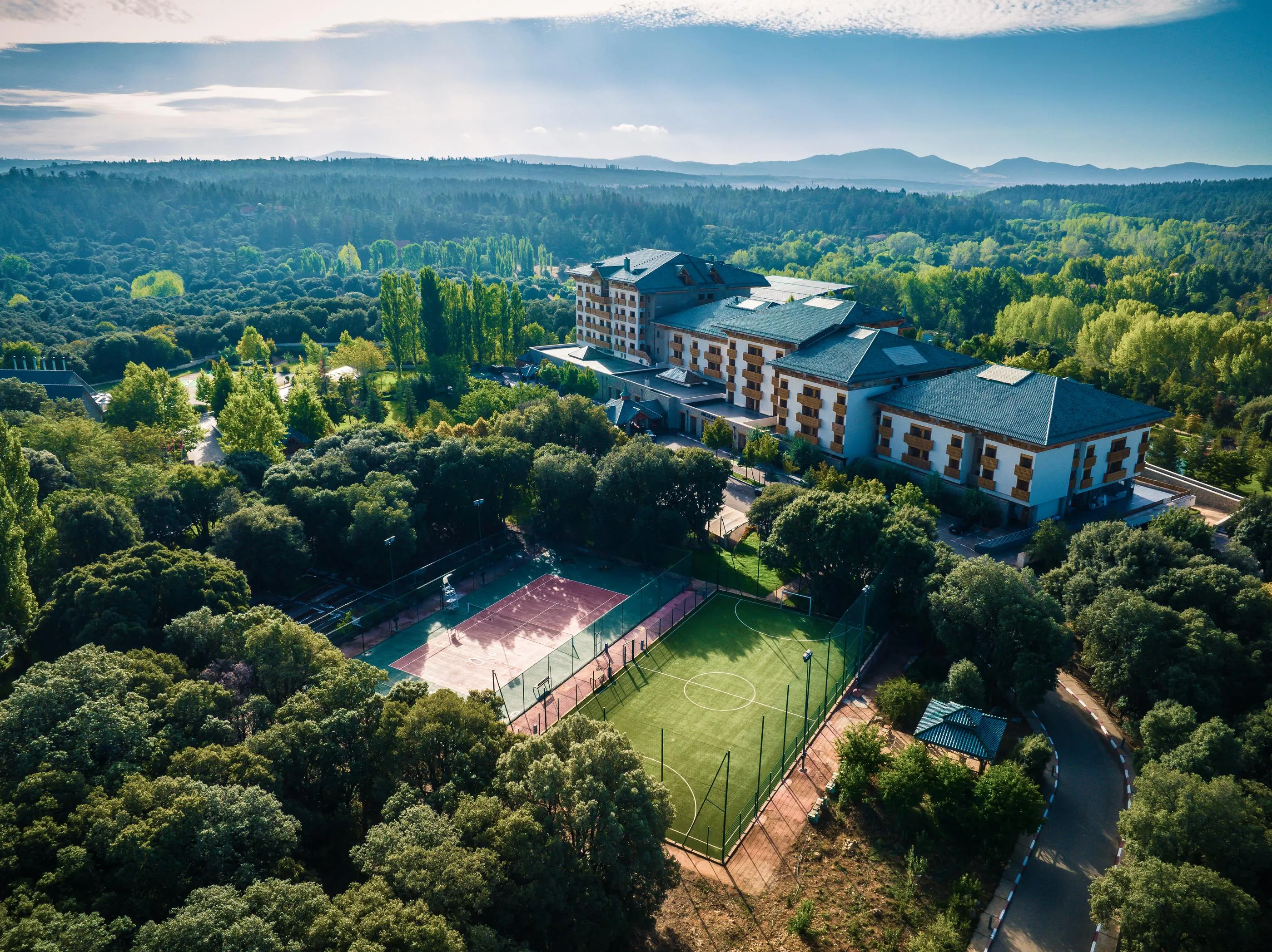 Aerial view of a building surrounded by trees with tennis courts and a sports field below, in a forested and mountainous landscape.