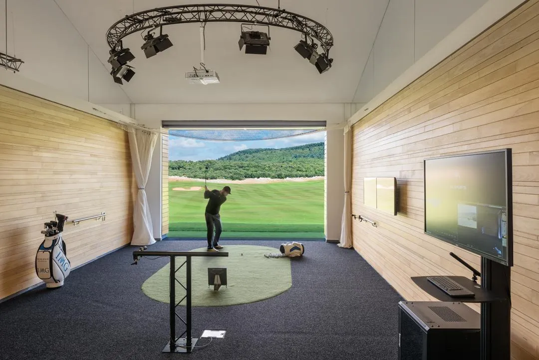 A golfer practices in an indoor golf simulator with a screen projecting a course, equipment, and a monitor nearby.