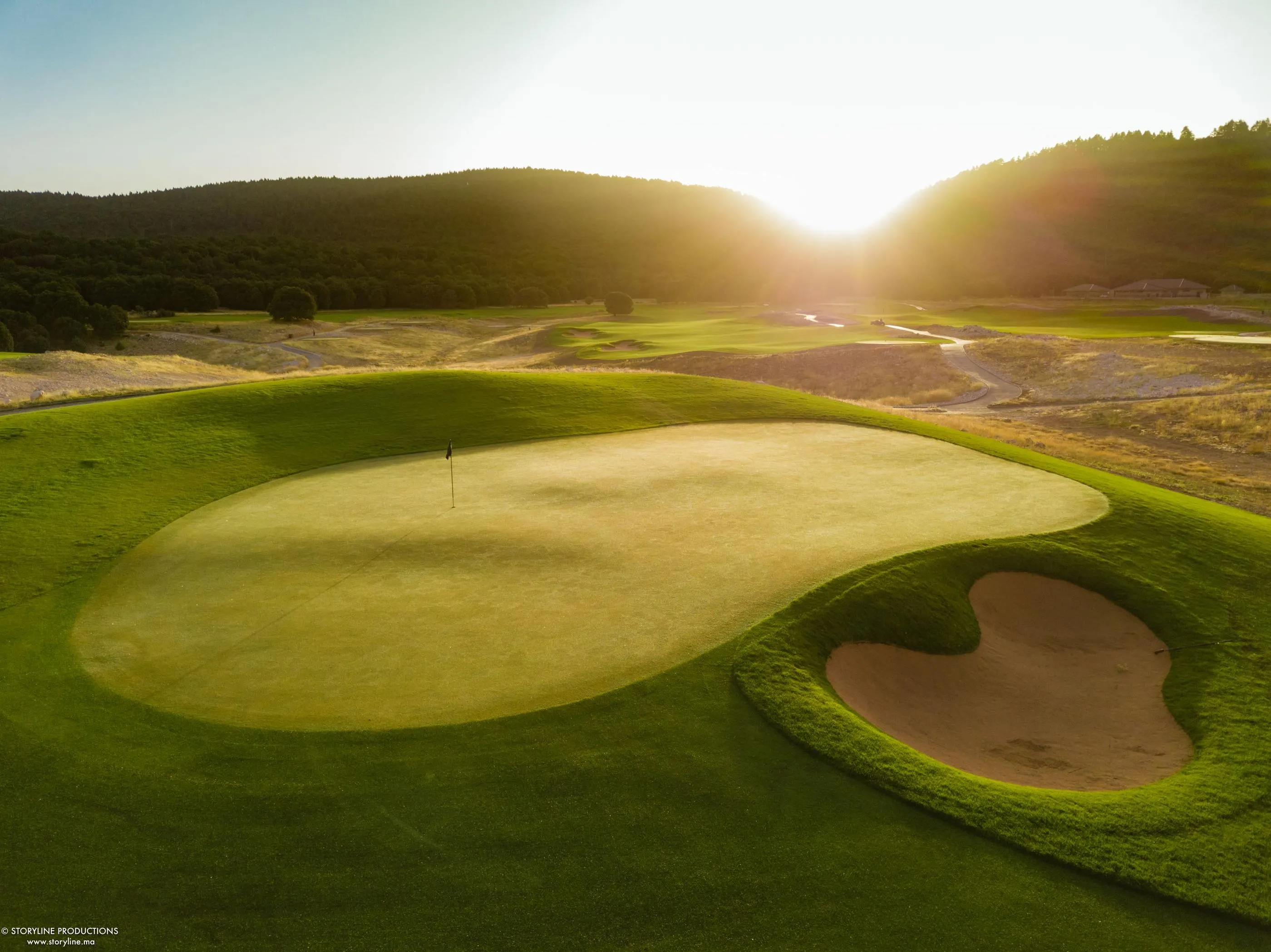 A golf green with a flag in the center, surrounded by sand bunkers, under a setting sun with wooded hills in the background