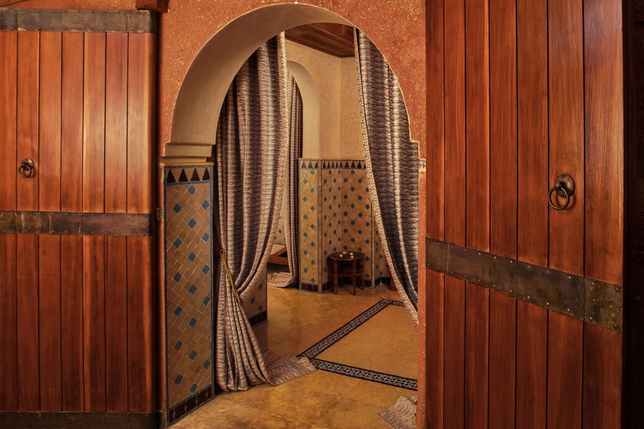 Two wooden doors with metal rings open onto a room with tiled walls and arches draped in fabric.