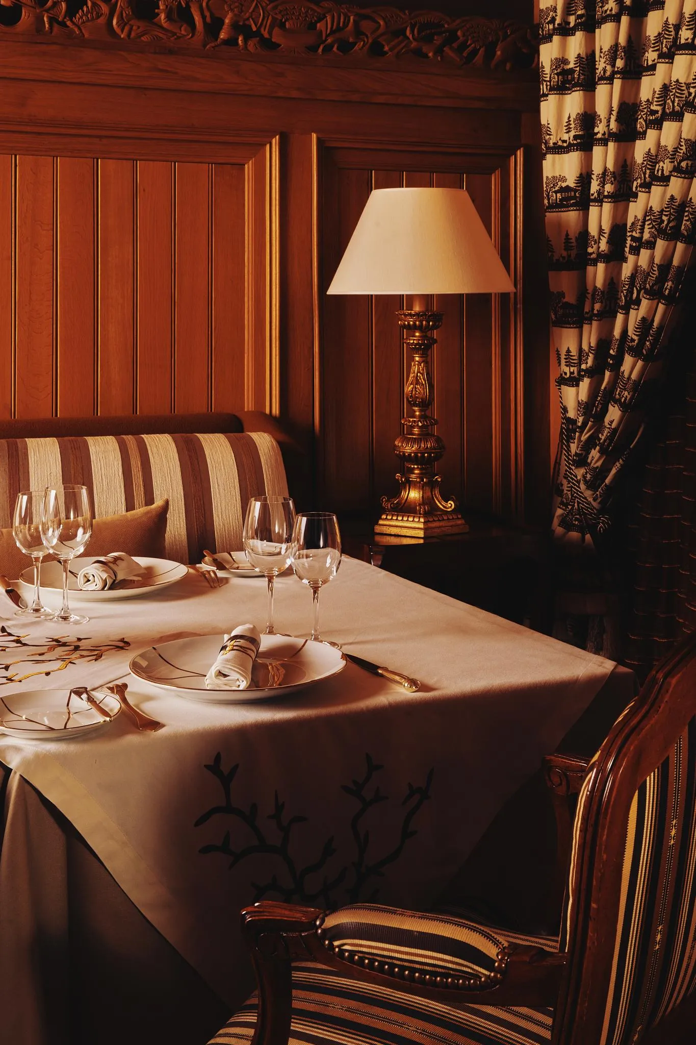 Table dressée avec assiettes, verres et couverts, éclairée par une lampe, dans un cadre en bois avec des rideaux à motifs
