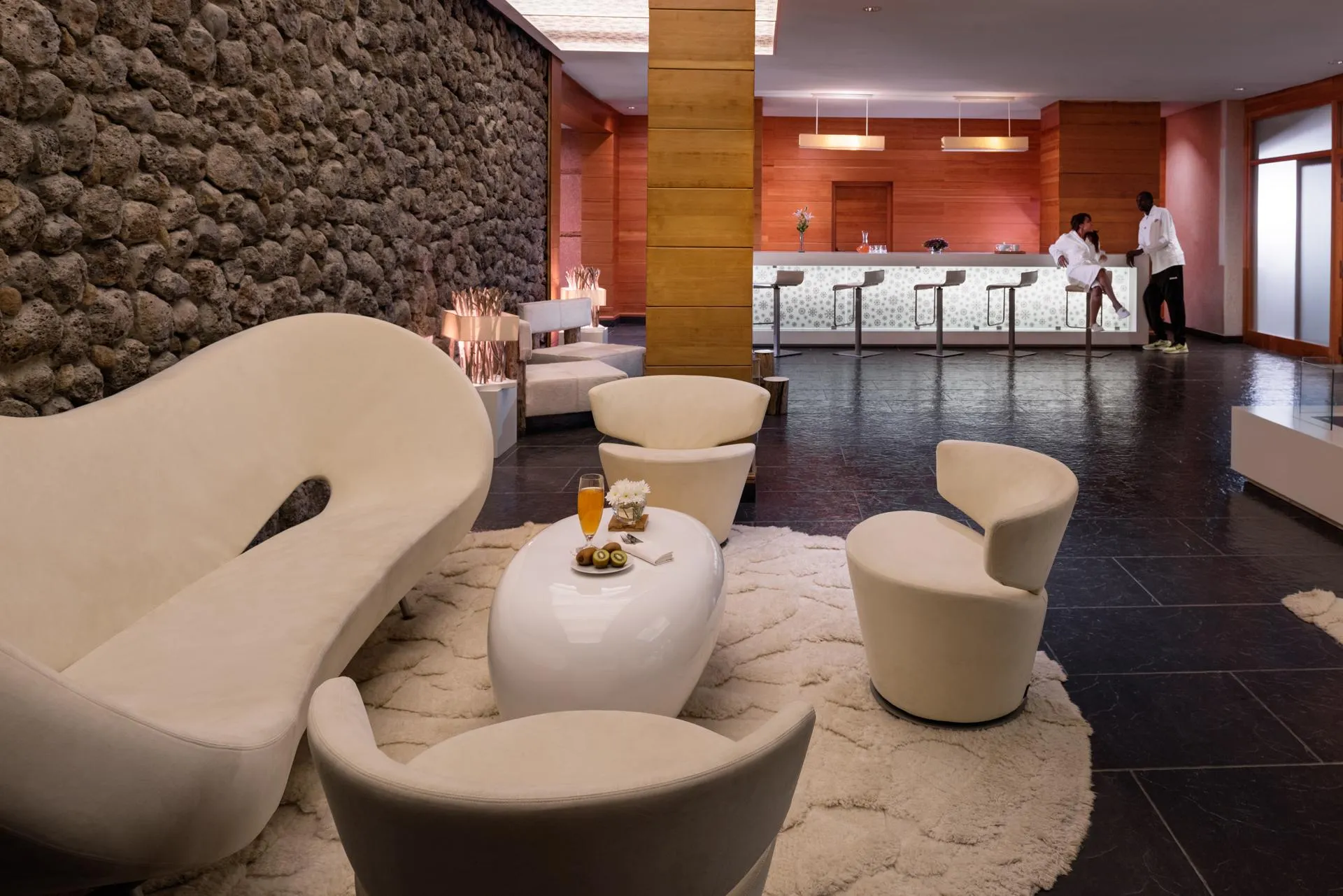 A hotel lounge with modern seating, a coffee table with drinks and fruit, and a bar with stools in the background.