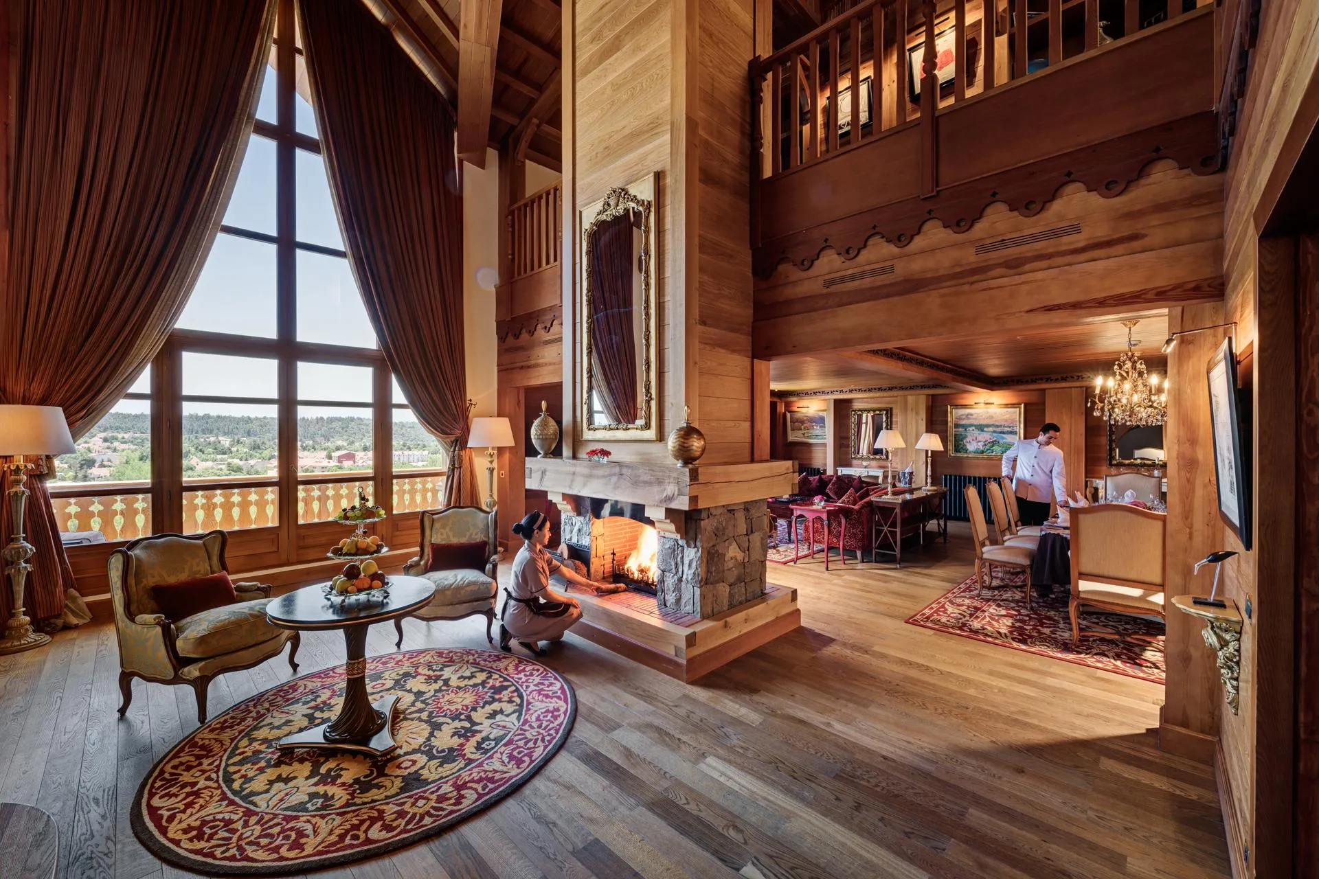 A wooden living room with a large window, a lit fireplace, armchairs, a coffee table, and people near the fire and the dining table.