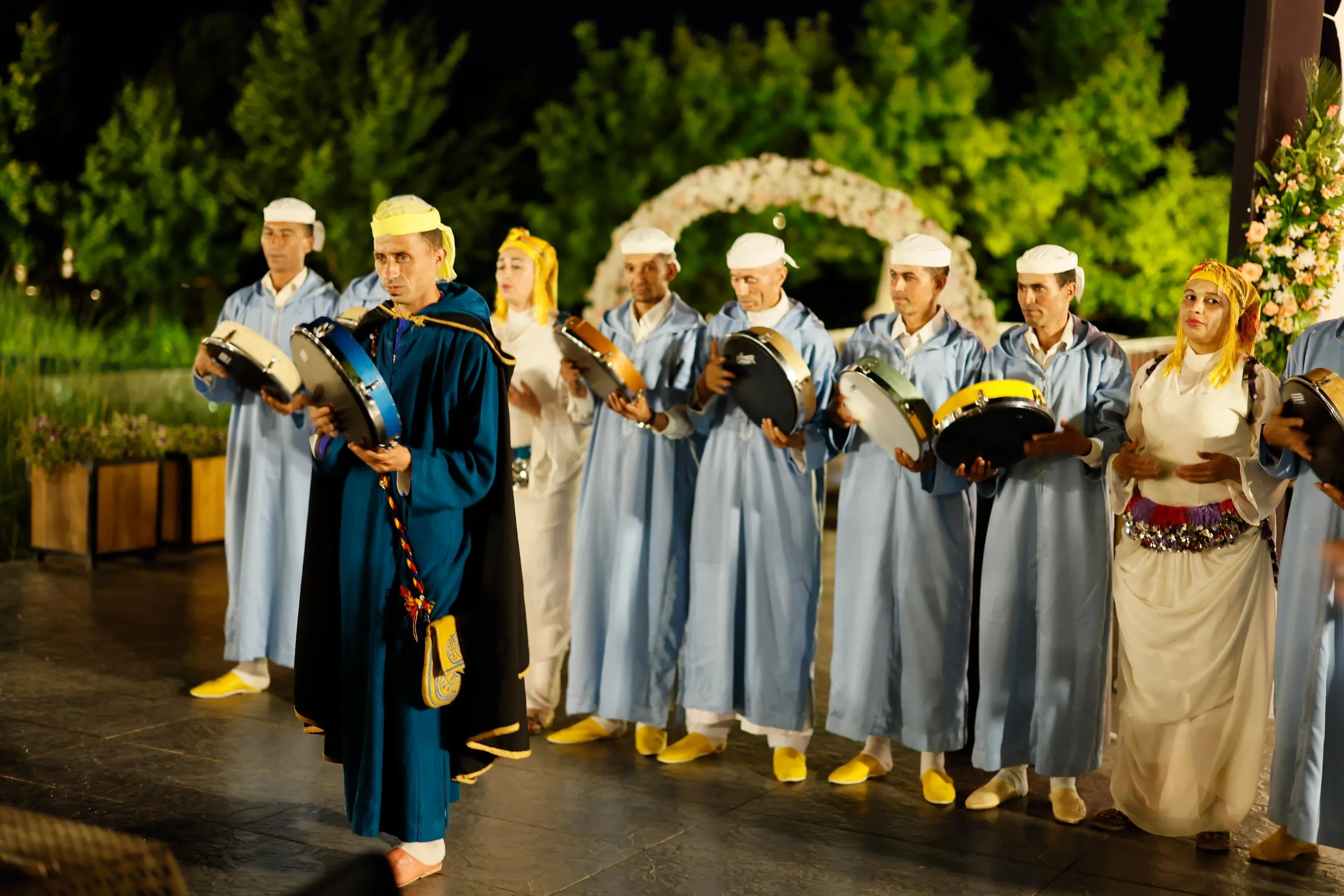 A group of people in traditional costumes plays small drums on an outdoor stage decorated with flowers.