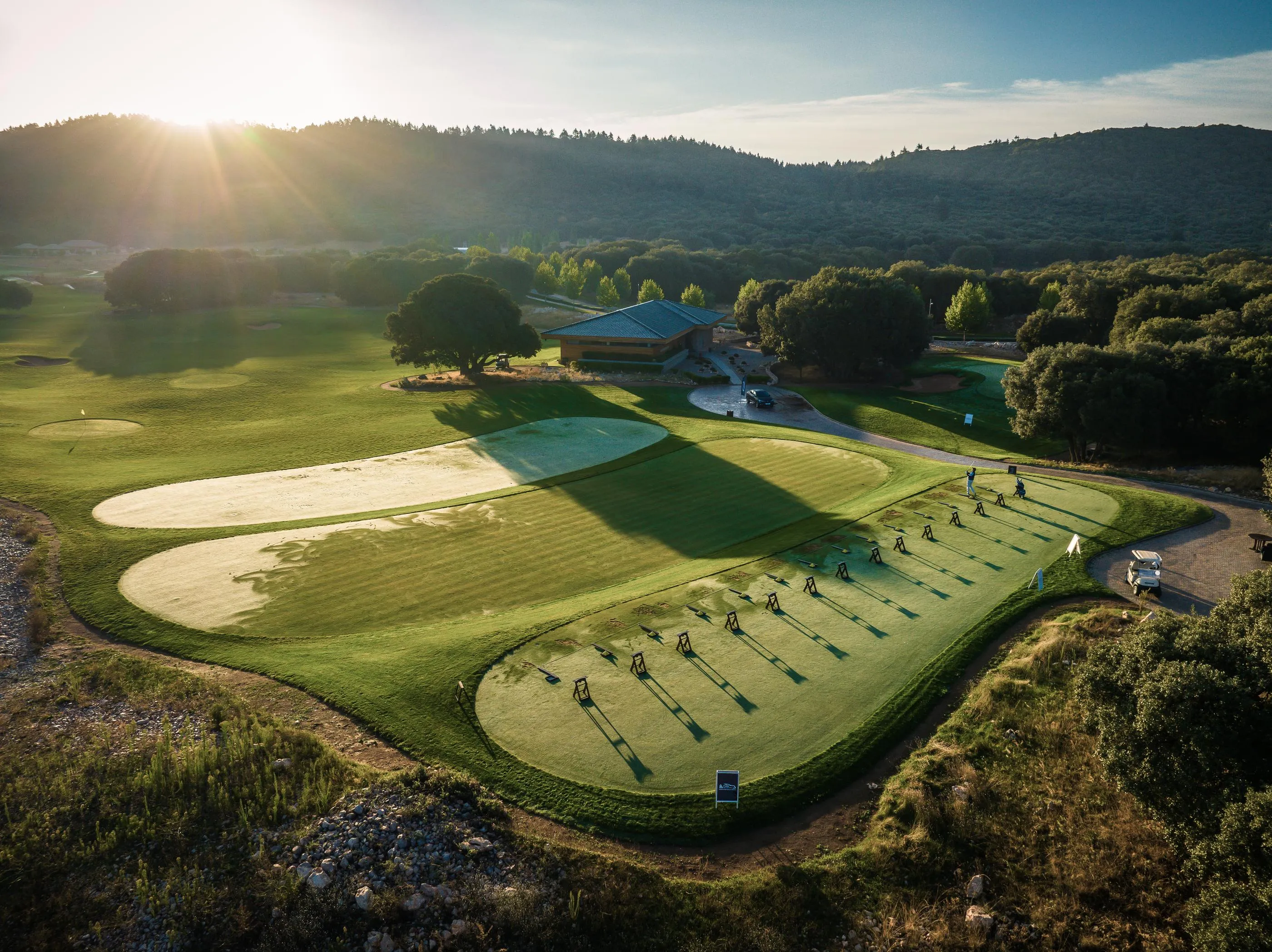 Aerial view of a golf course with players in action, sand bunkers, and a clubhouse in the background under a clear sky.