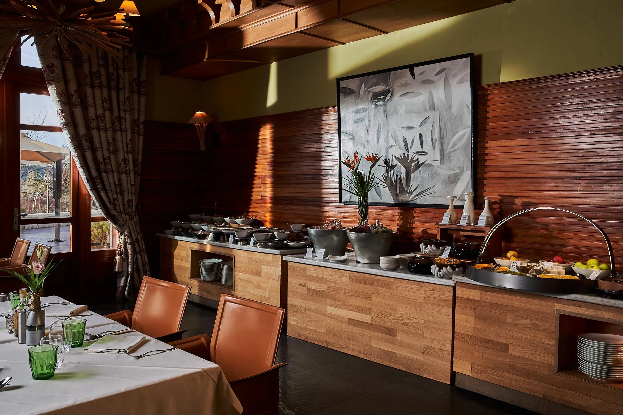 A breakfast buffet with fruits, drinks, and hot dishes in a restaurant with wooden walls and large windows.