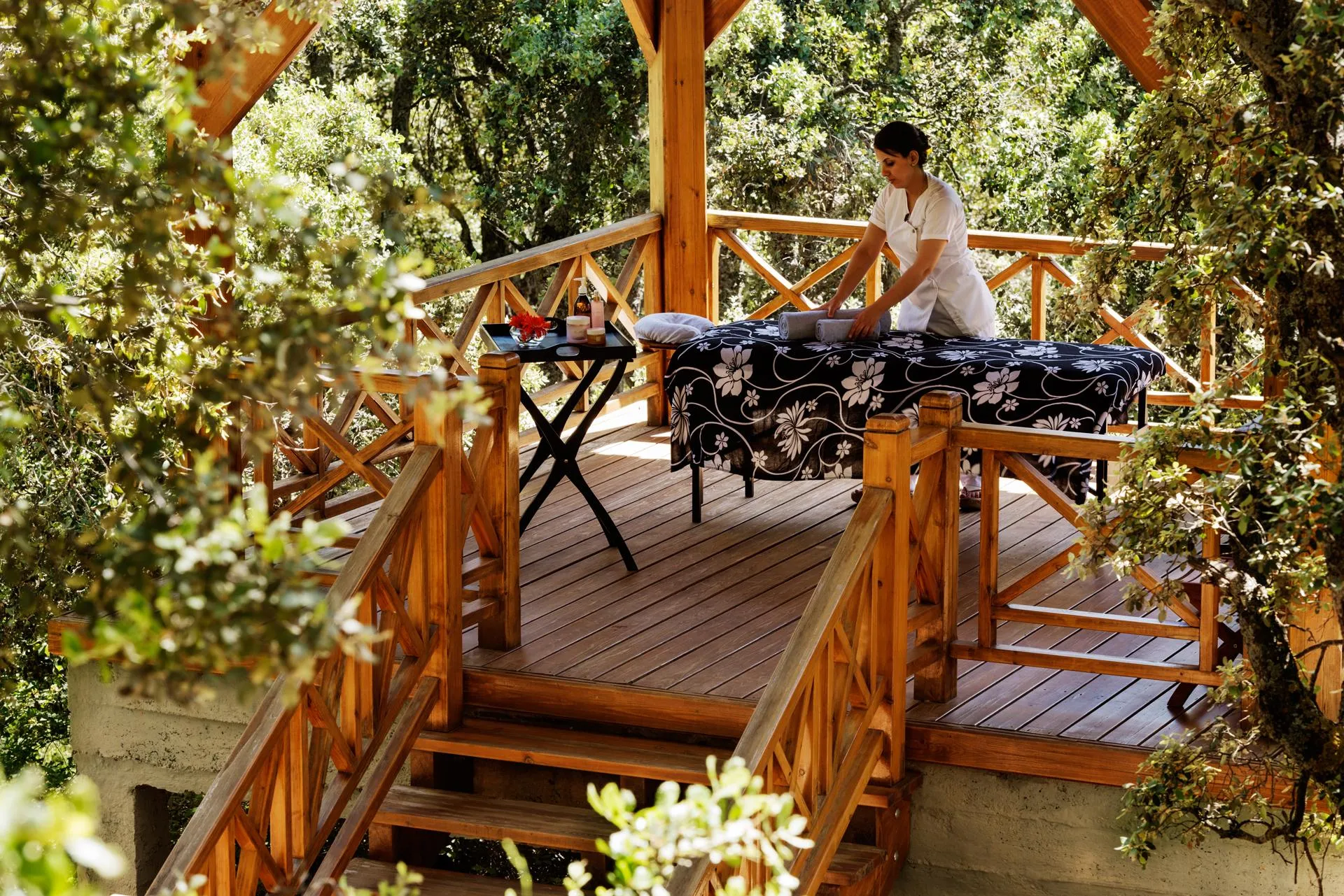 A person in white clothing is giving a massage to a person lying on a table outside, surrounded by trees.