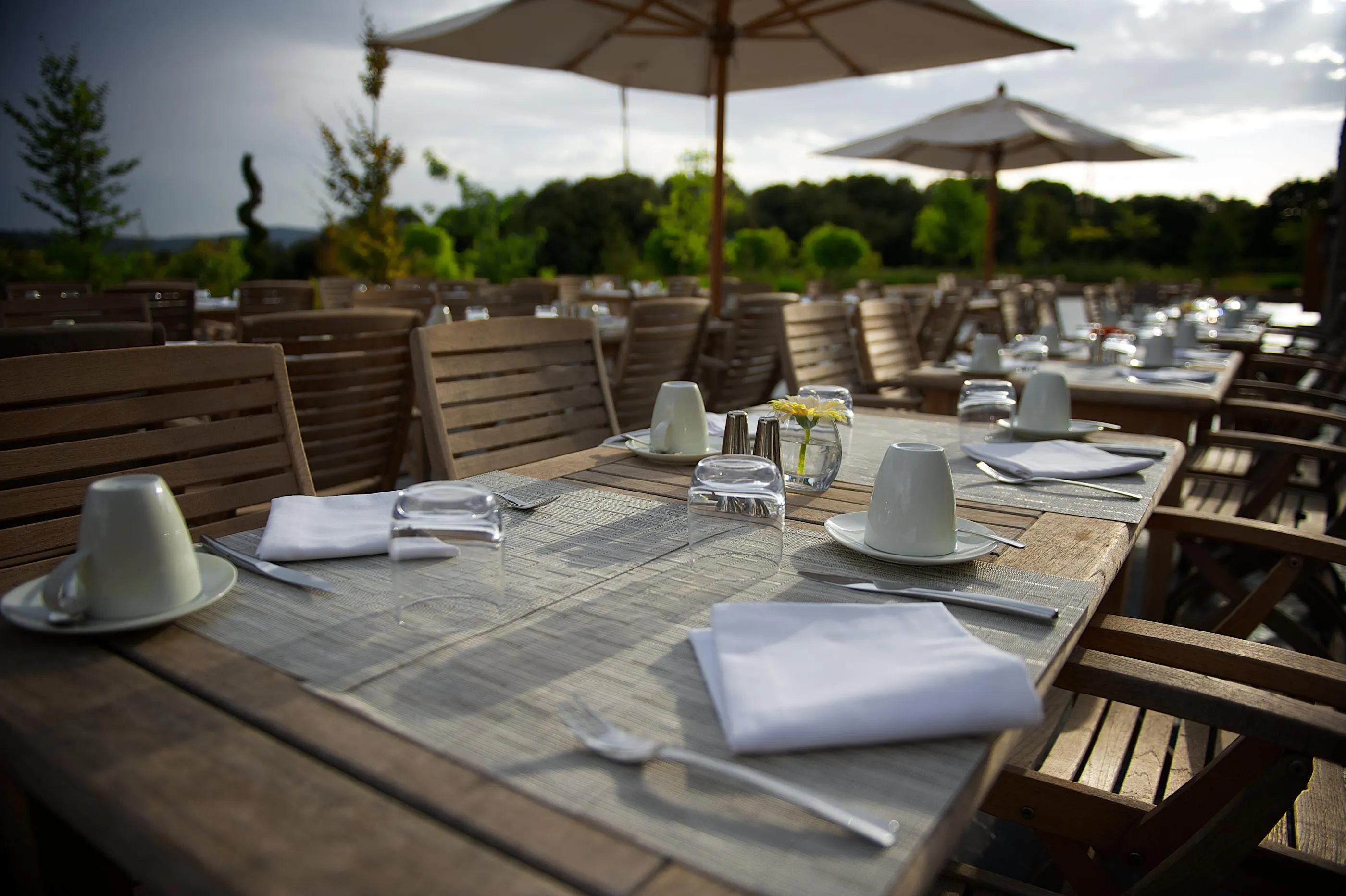 Outdoor terrace table with dishes, glasses, and napkins, surrounded by wooden chairs and shaded by umbrellas.