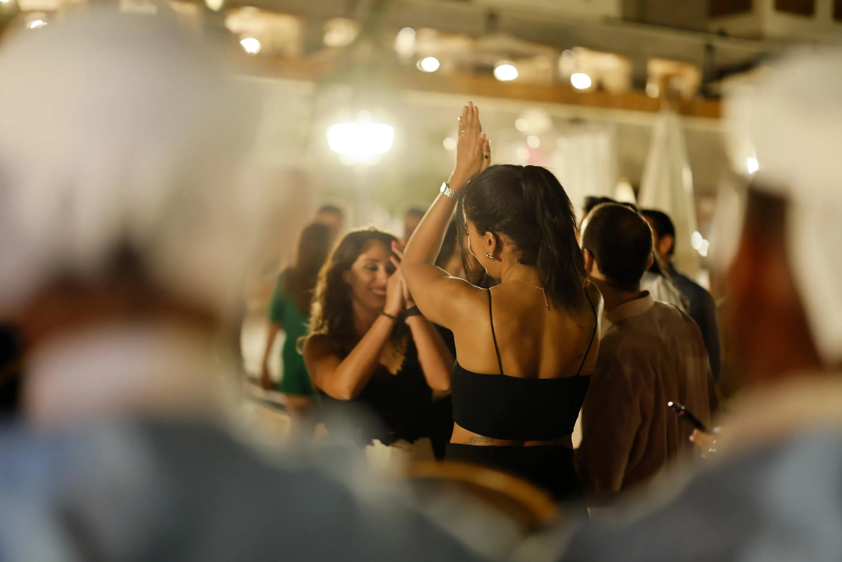 Two women are dancing together in a lit room, surrounded by other people socializing.