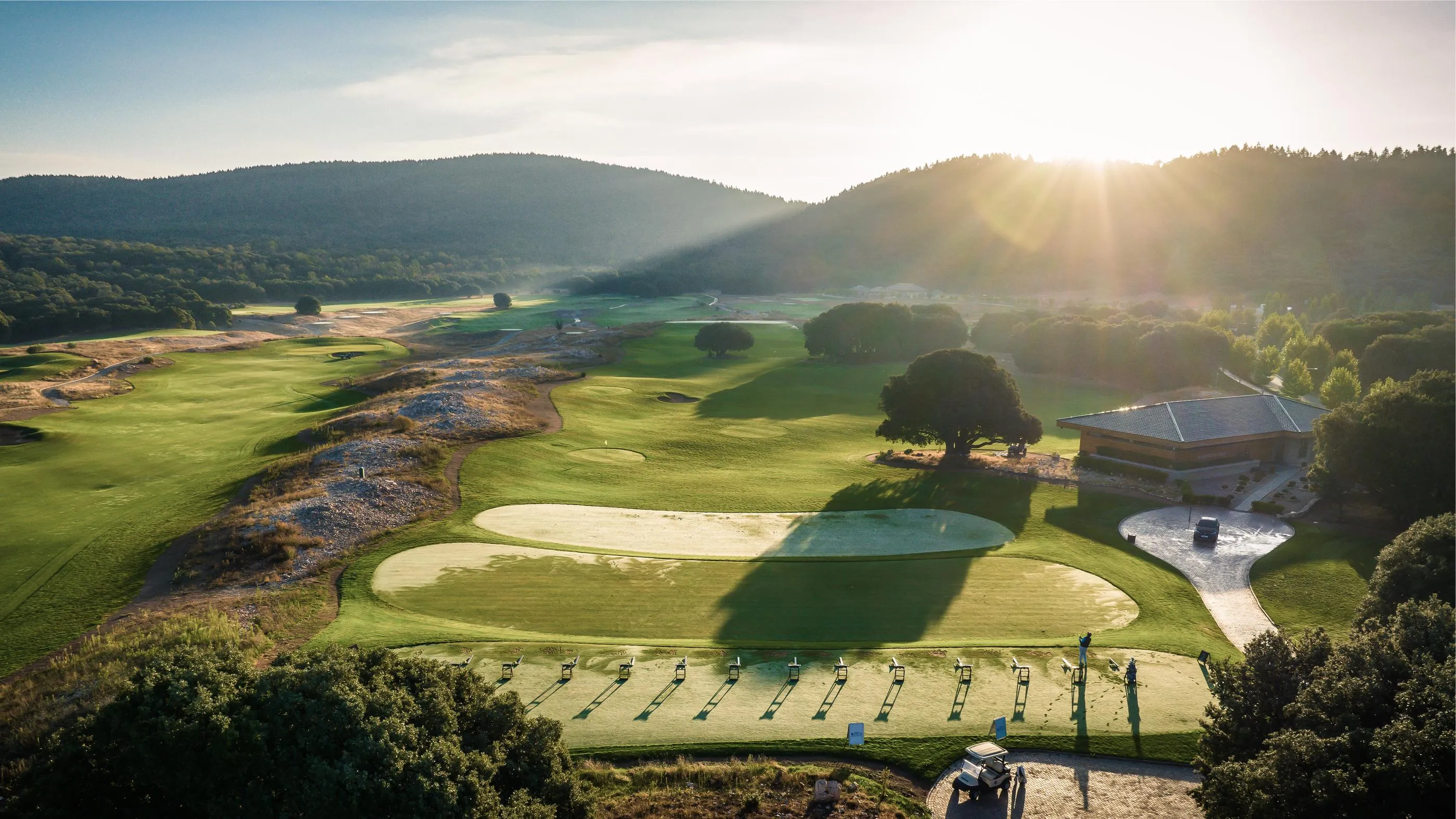 A golf course with practice greens and targets, surrounded by wooded hills, under a rising sun