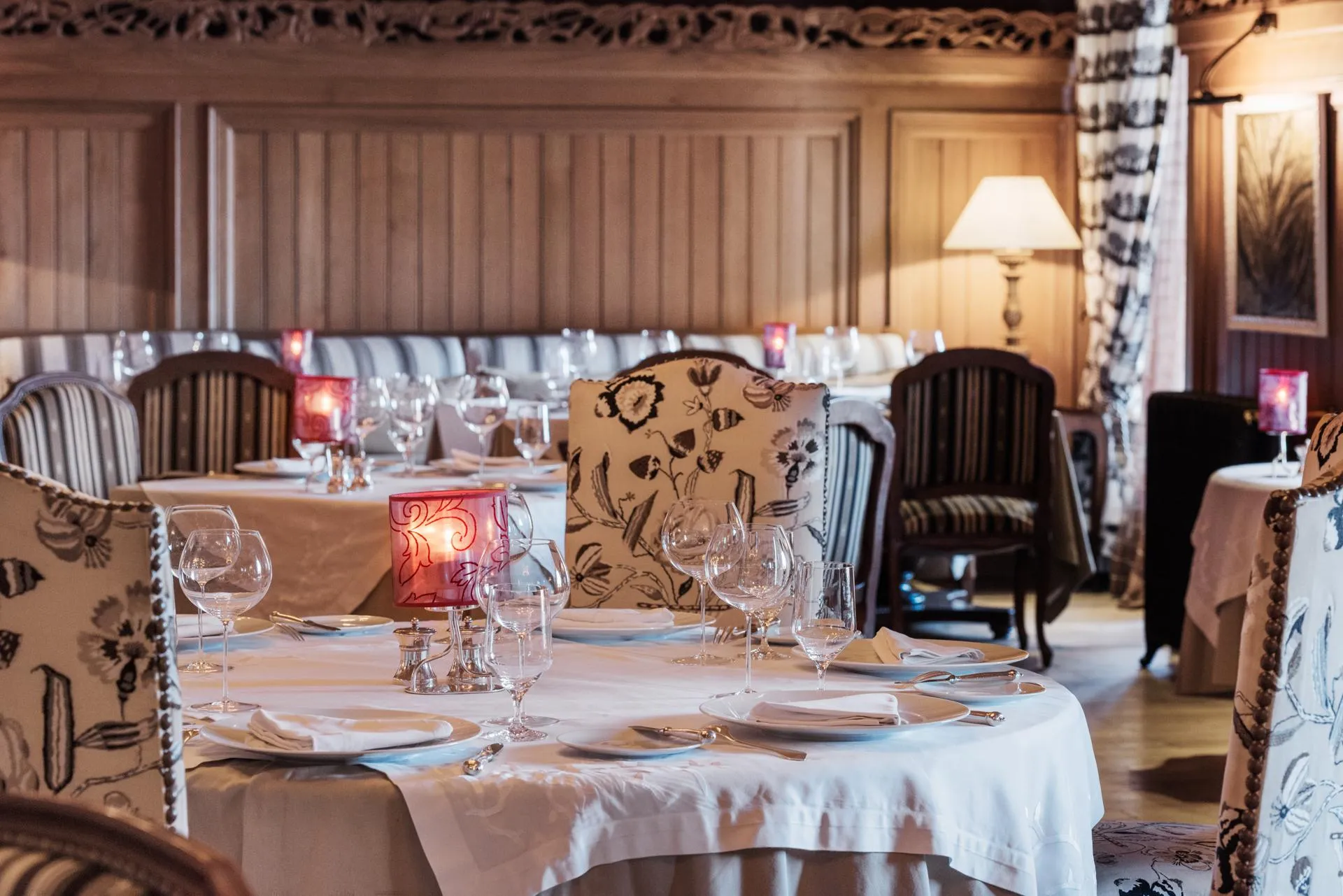A table set in a restaurant with glasses, cutlery, and candles, surrounded by upholstered chairs and decorated with tablecloths and floral-patterned curtains.
