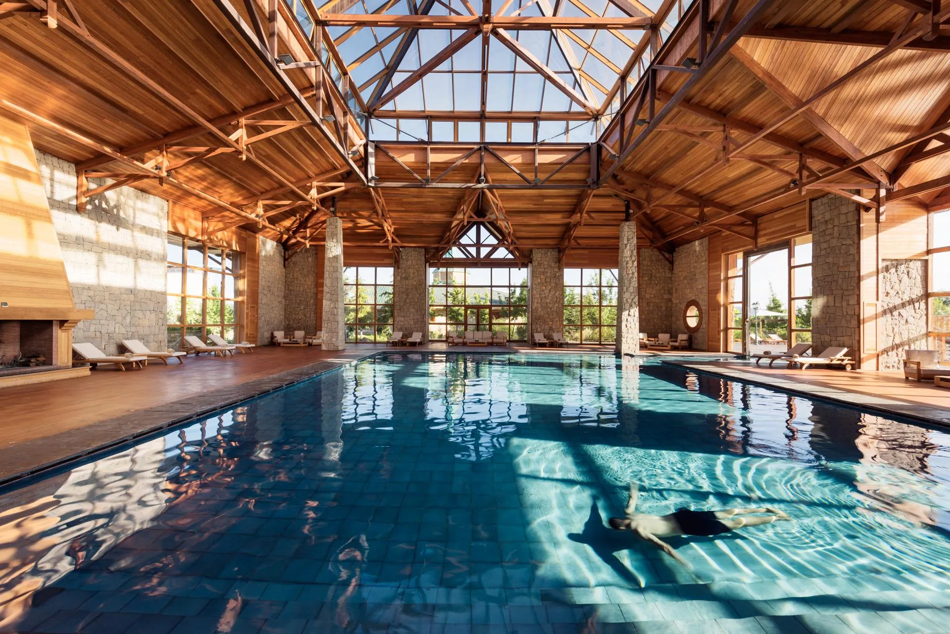 Indoor pool with a wooden roof and large windows, surrounded by lounge chairs and stone columns