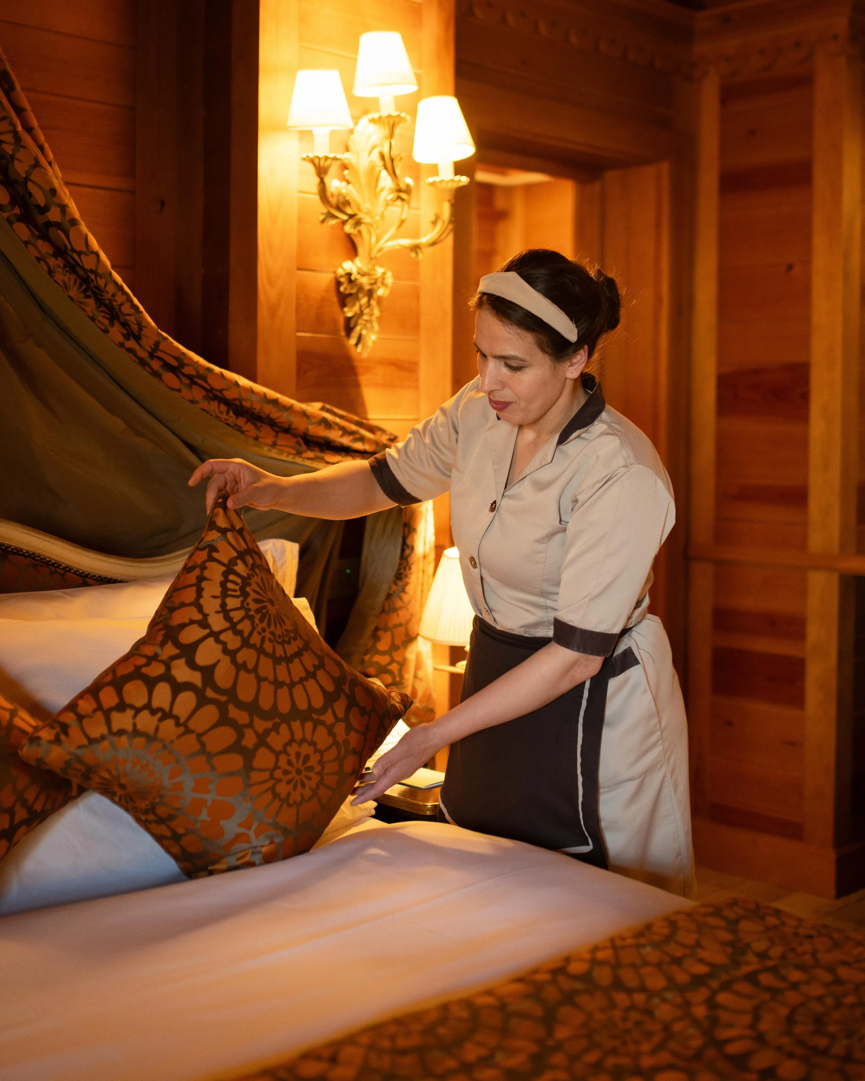 A woman in a maid's uniform arranges cushions on a bed in a room with antique decor.