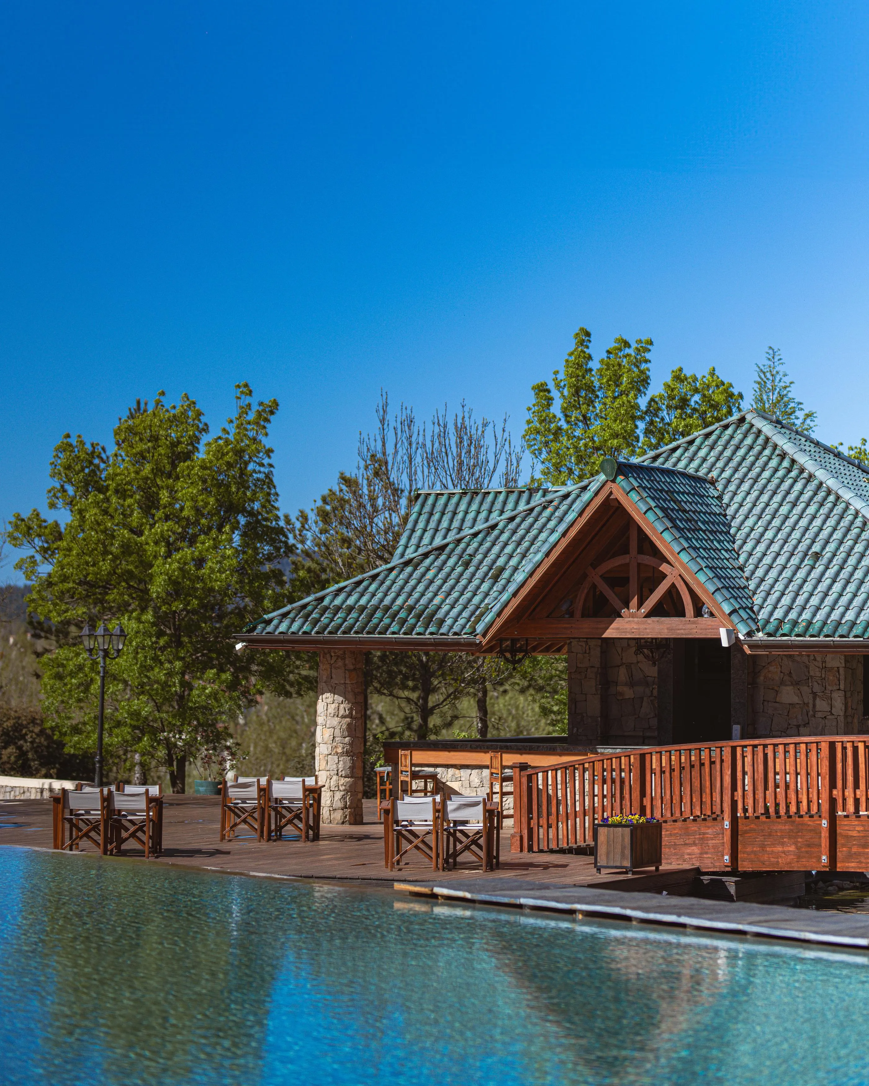 A wooden pavilion with a green-tiled roof near a pool surrounded by lounge chairs.