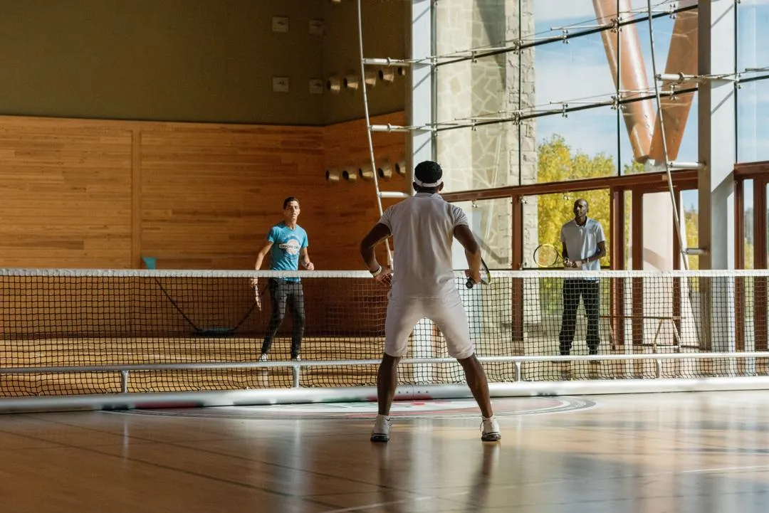 Three people are playing table tennis in a room with wooden walls and large windows.