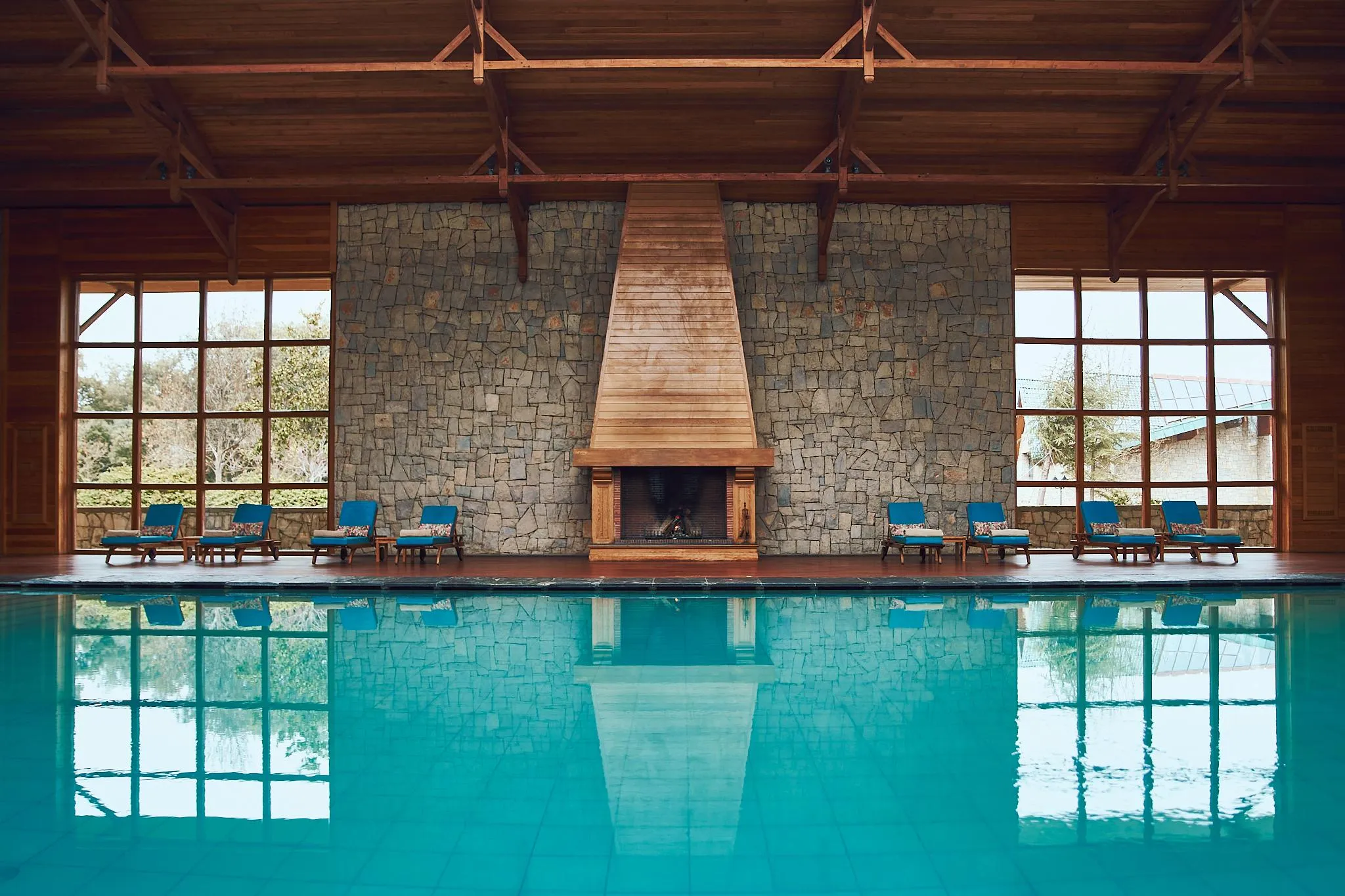 Indoor pool with sun loungers in front of a stone wall and a lit fireplace, surrounded by large wooden windows