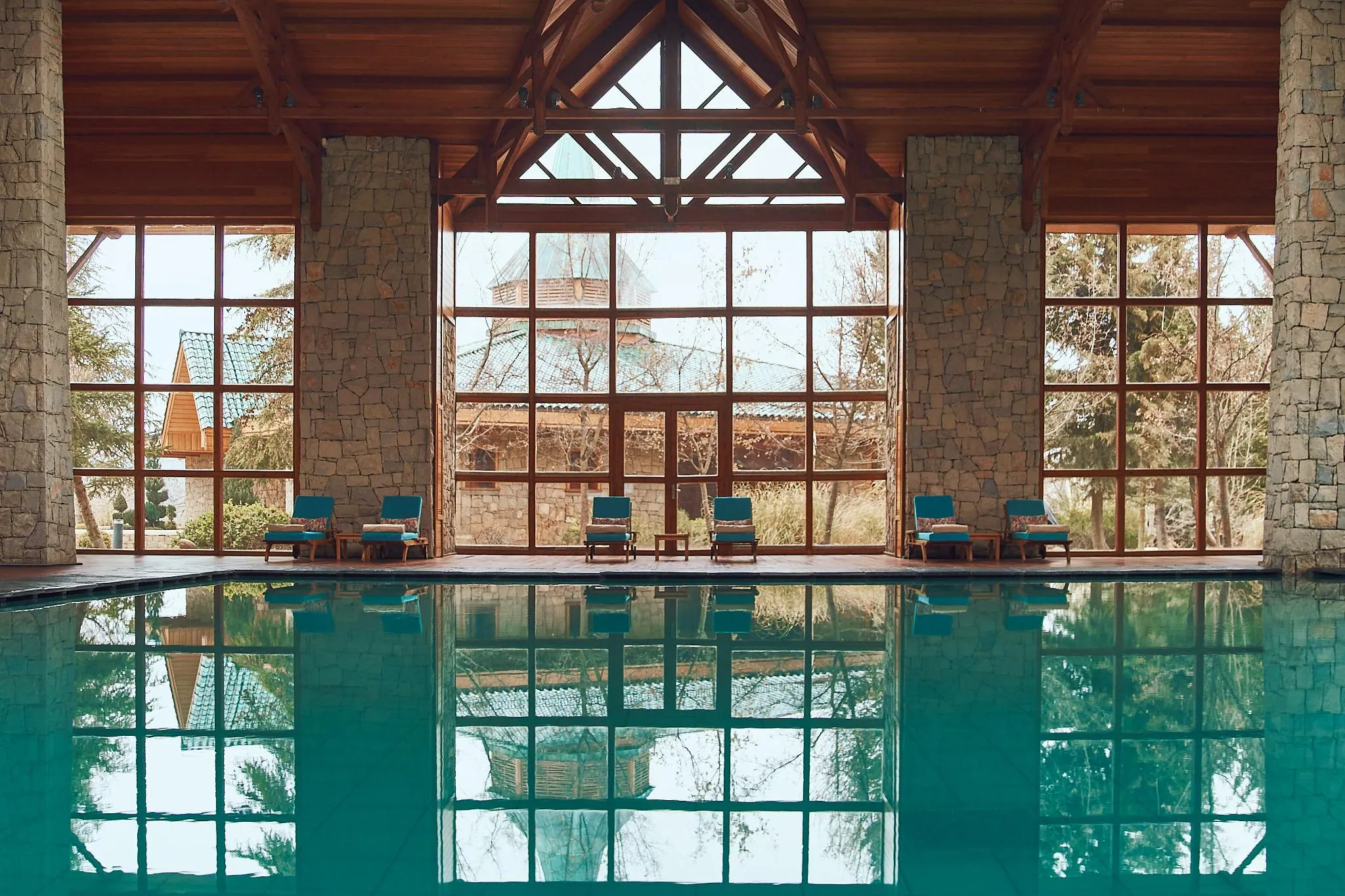 Indoor pool with a view of lounge chairs and stone and wood structures through large windows