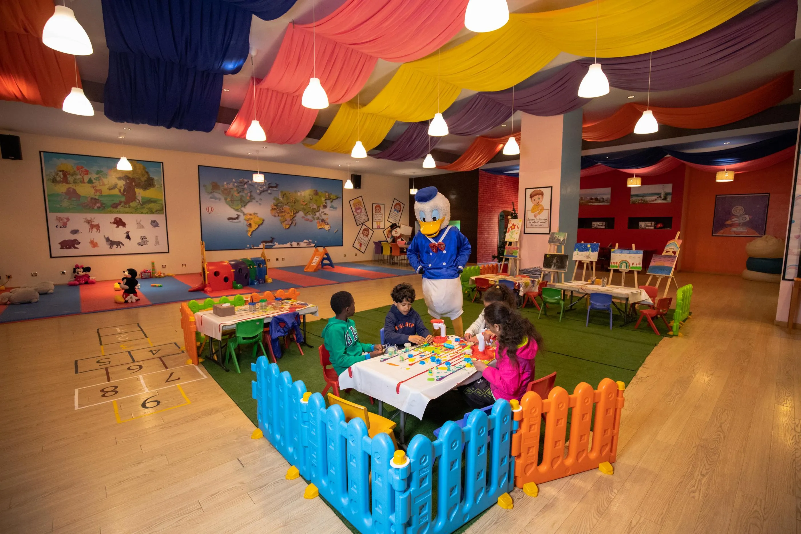 A colorful playroom with children seated at tables, a cartoon character in costume, and educational wall decorations.