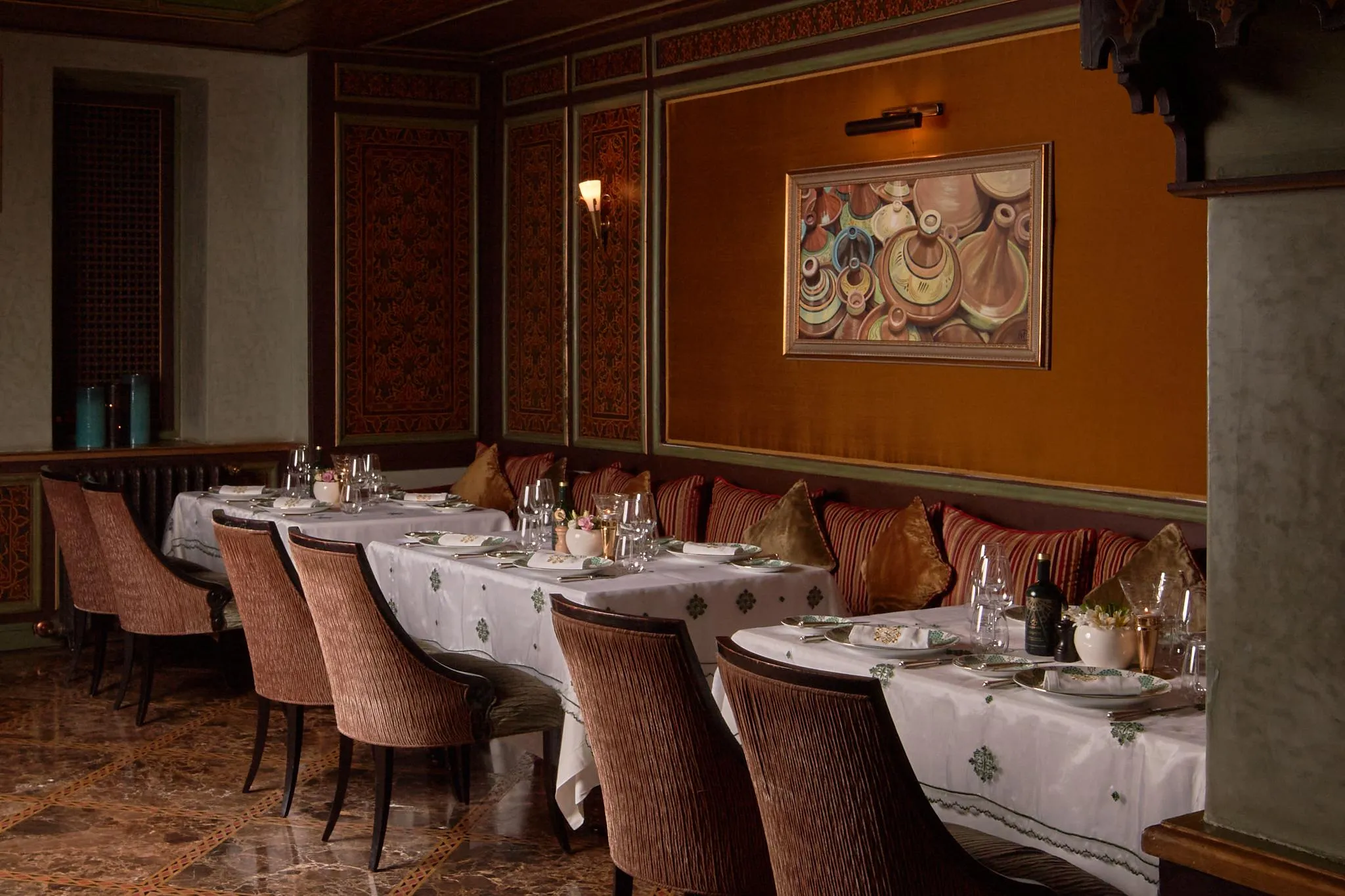 An elegant dining room with set tables and upholstered chairs, decorated with cushions and a colorful wall painting.