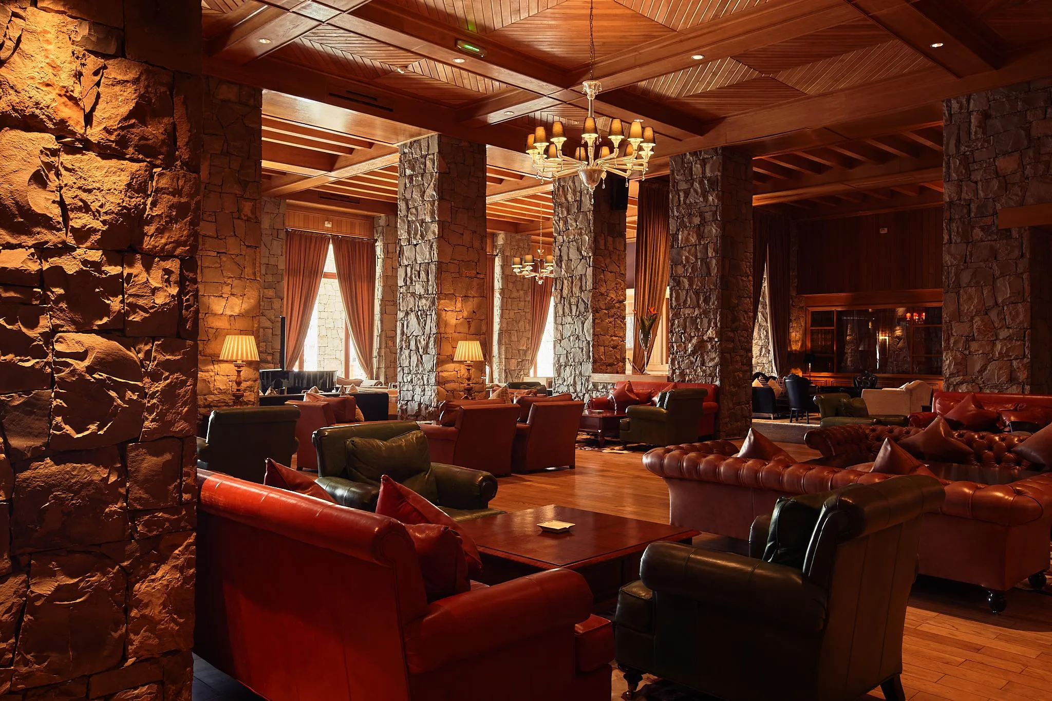 A living room with brown and green leather sofas, lamps, and chandeliers, surrounded by stone walls and with a wooden ceiling.