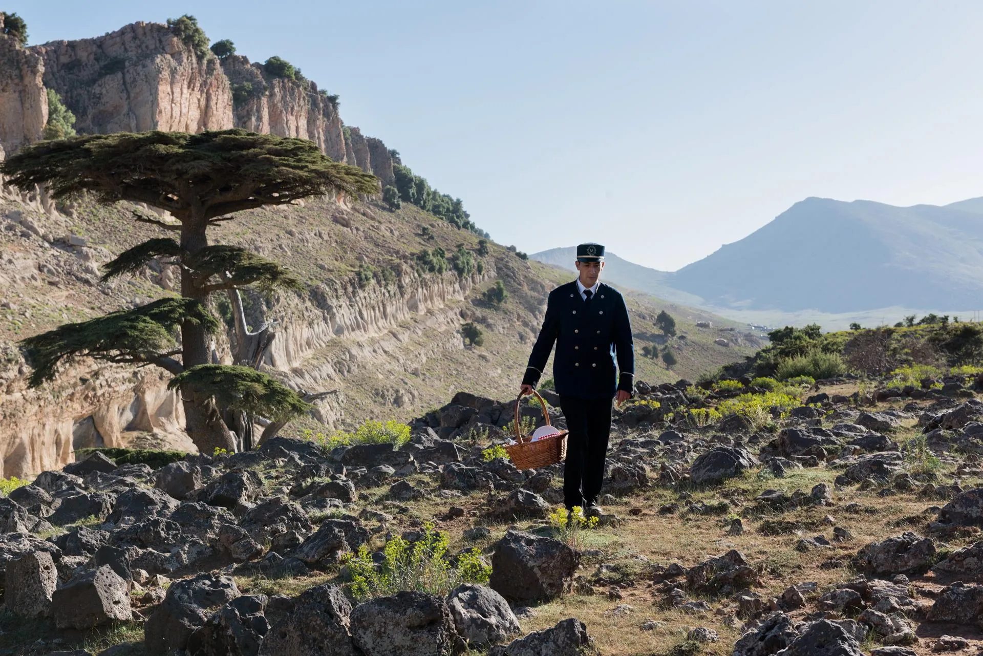 A man in uniform walks through a rocky mountainous landscape holding a basket.