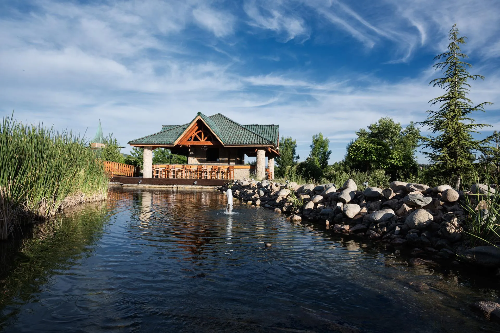 A wooden pavilion with a green roof located at the edge of a body of water surrounded by rocks and vegetation.