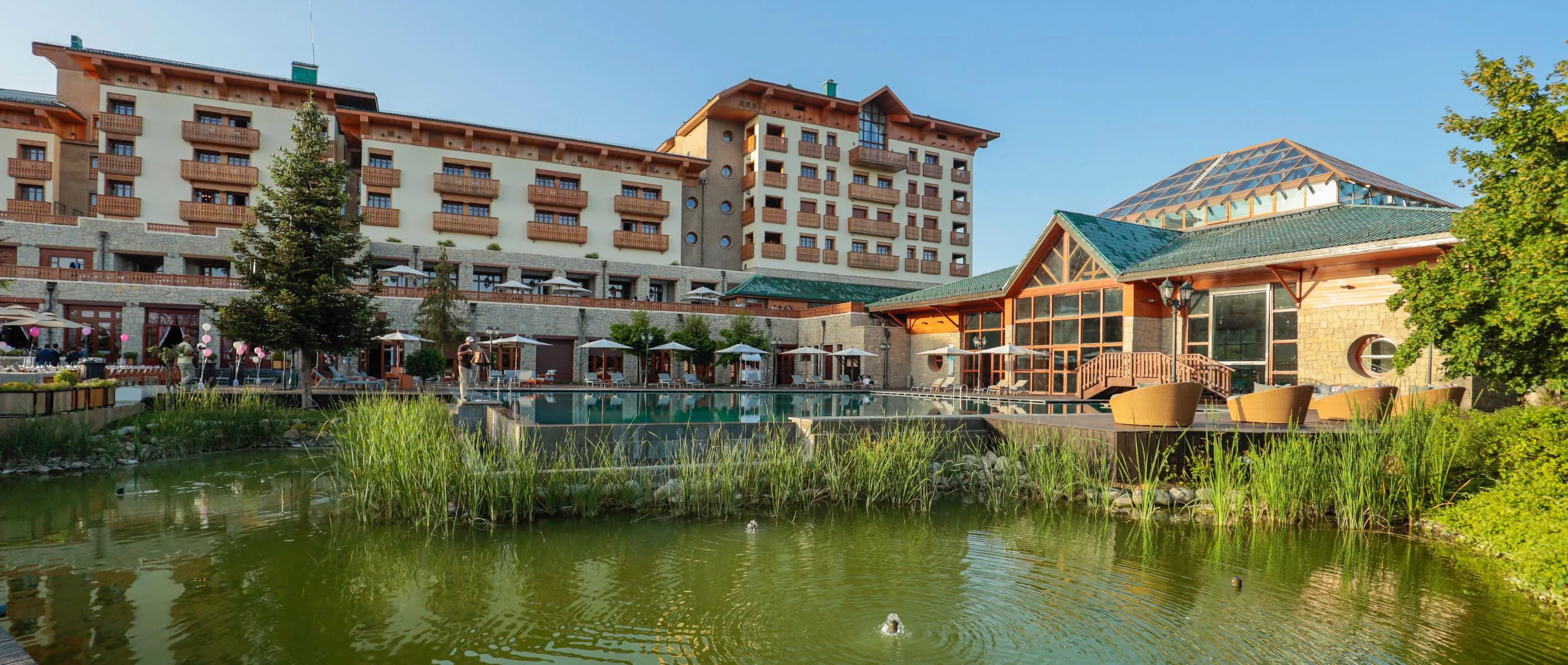 A hotel complex with stone and wood buildings, terraces with tables and chairs, and a water feature with aquatic plants in the foreground.
