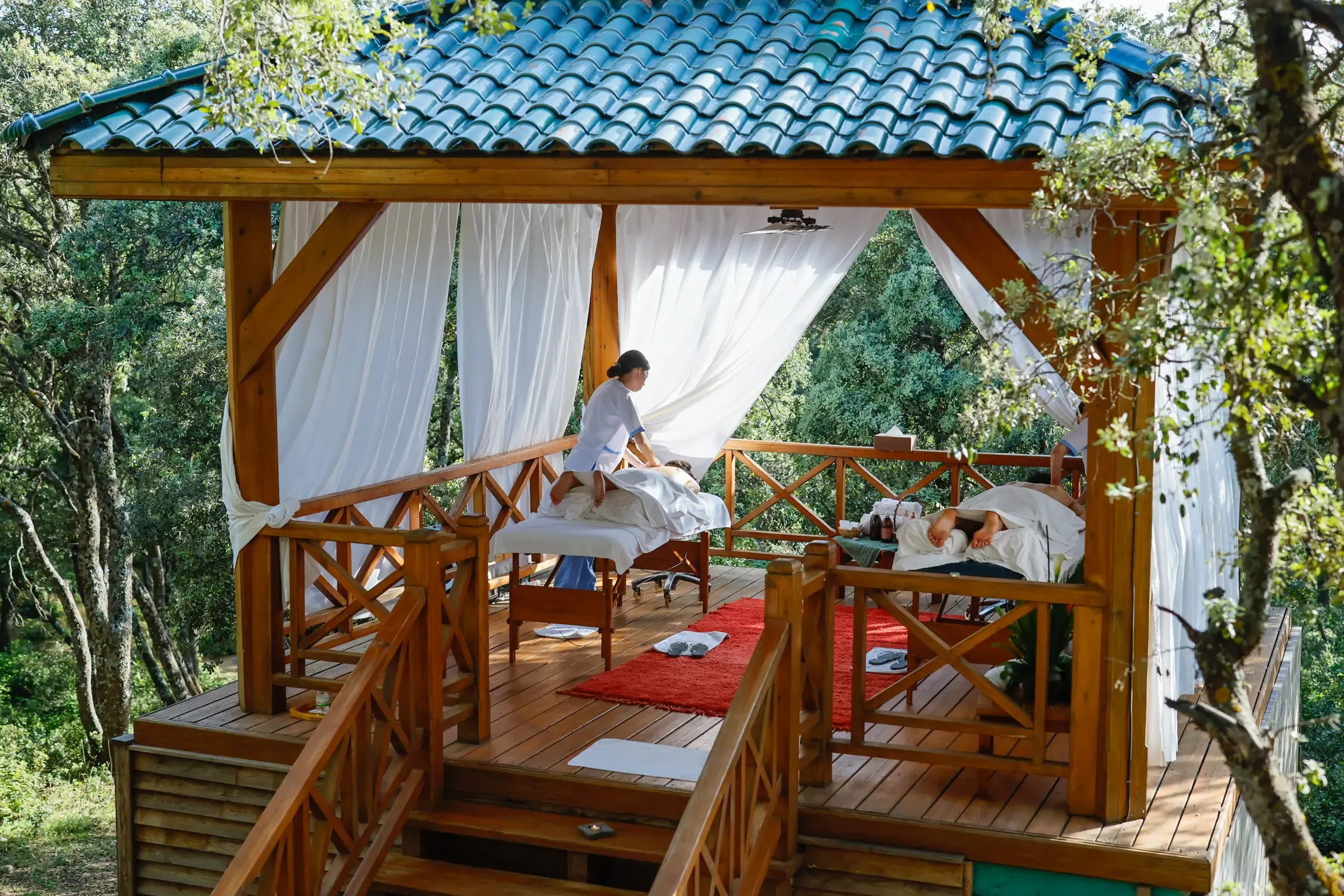 Deux personnes allongées sur des tables de massage en plein air sous un auvent en bois avec un toit en tuiles bleues, entourées d'arbres