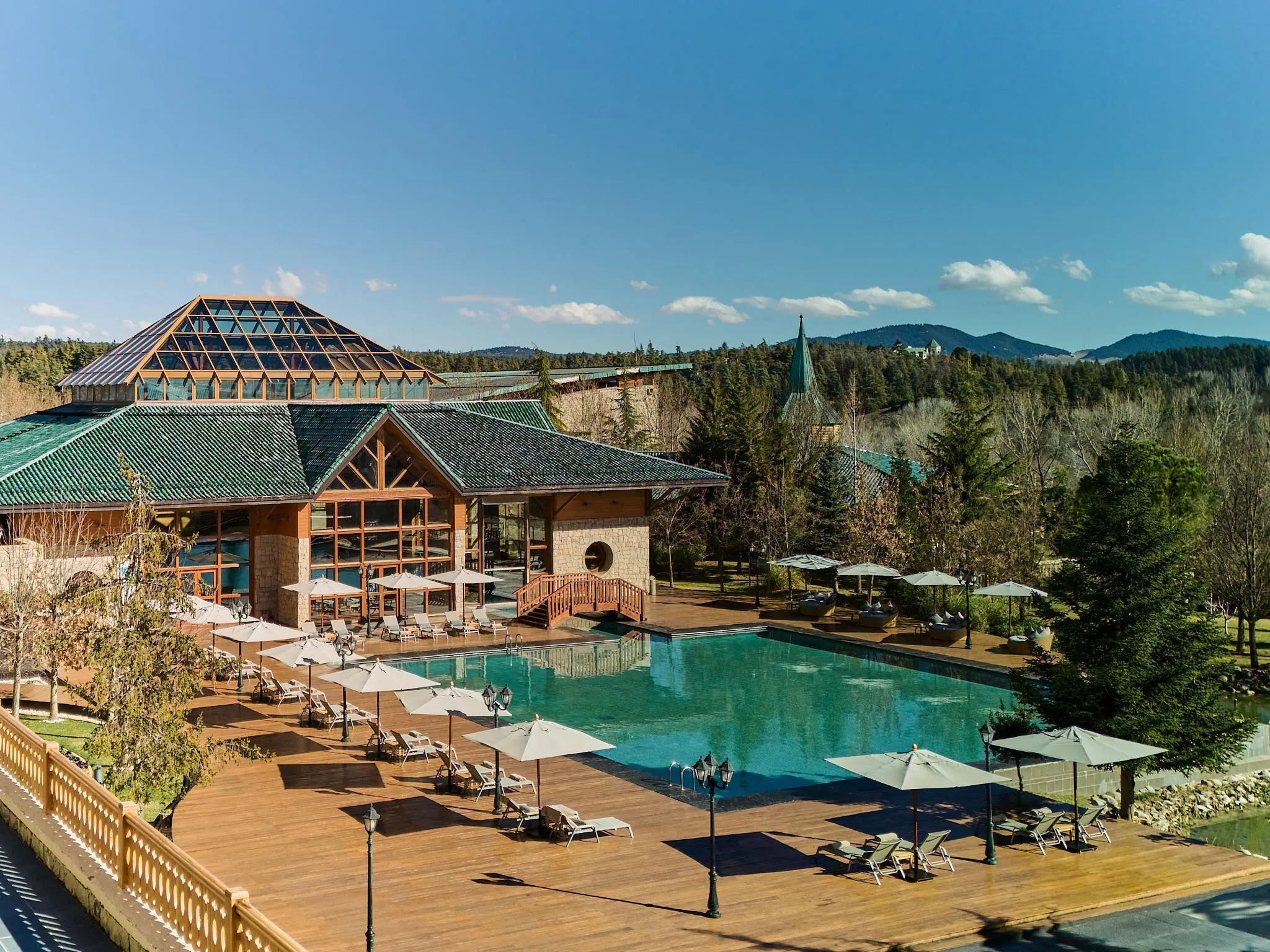 Outdoor swimming pool surrounded by sun loungers and parasols, with a wooden building and green roof in the background, in a mountainous natural setting.