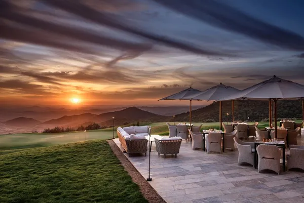 Terrace with chairs and tables under umbrellas, facing a sunset over mountains and a golf course.